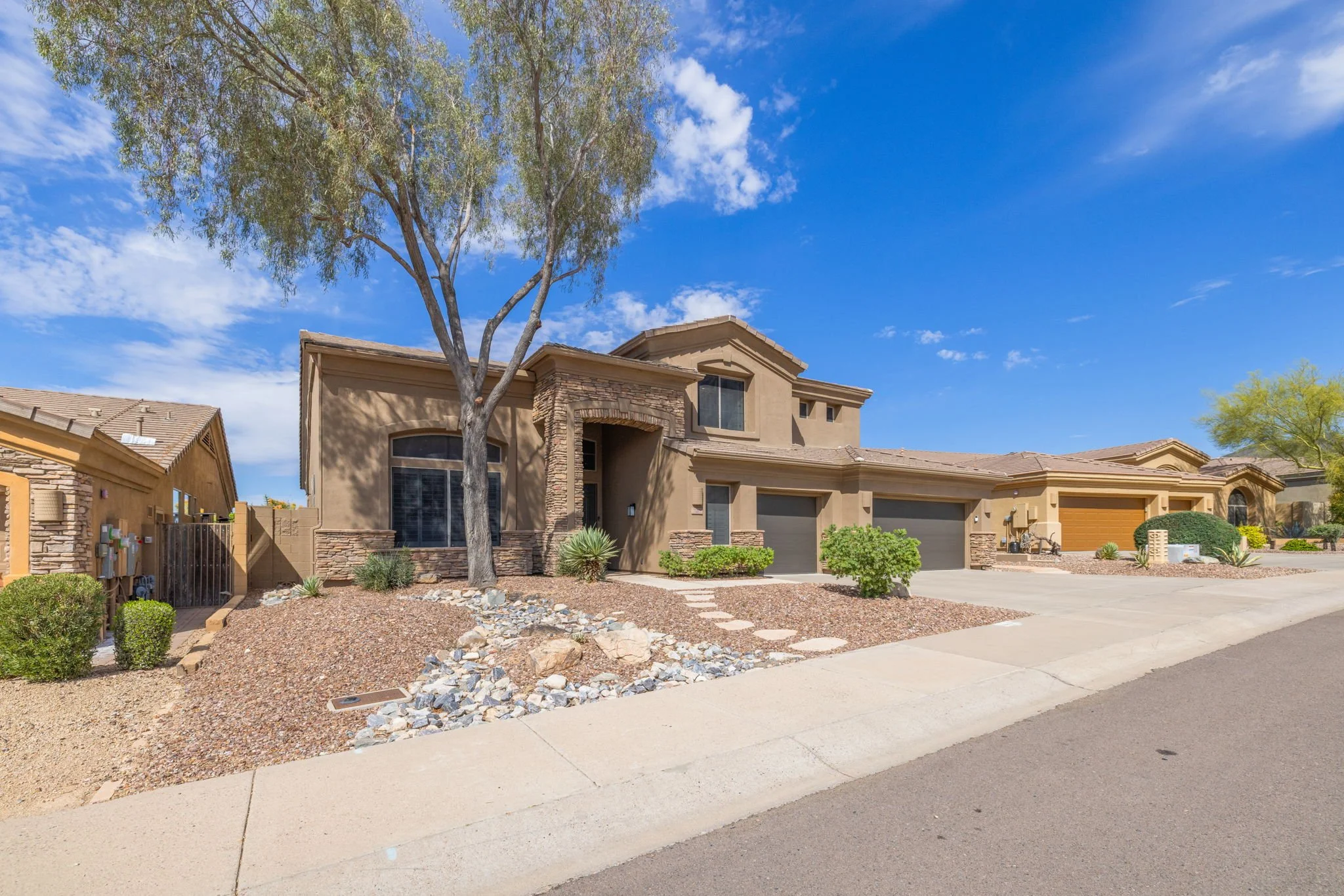 A modern suburban house with a tan exterior, stone accents, and a landscaped front yard featuring rocks, bushes, and a tree, under a bright blue sky with some clouds.