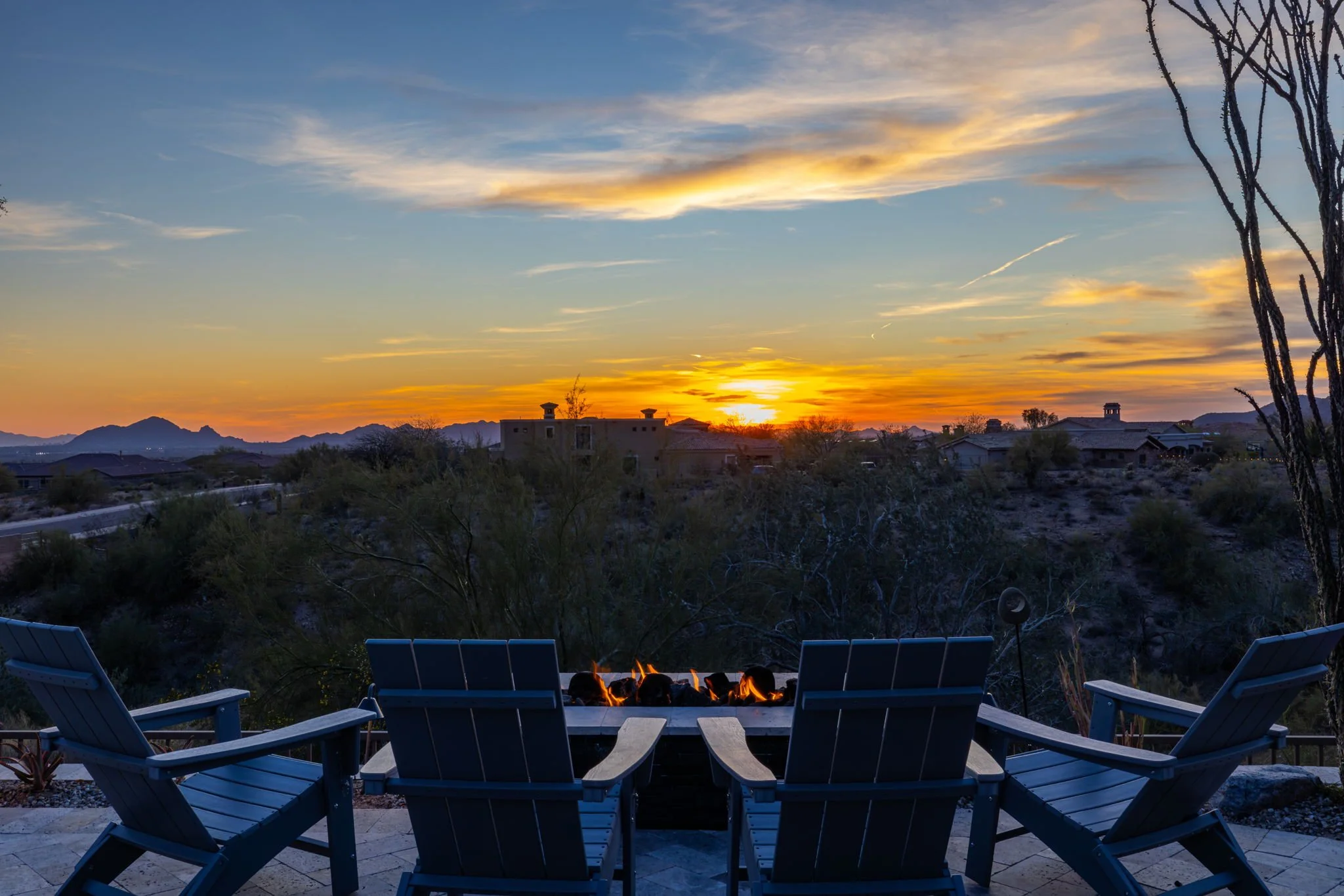 Sunset over desert landscape with distant mountains, viewed from a patio with four blue chairs and a small firepit.