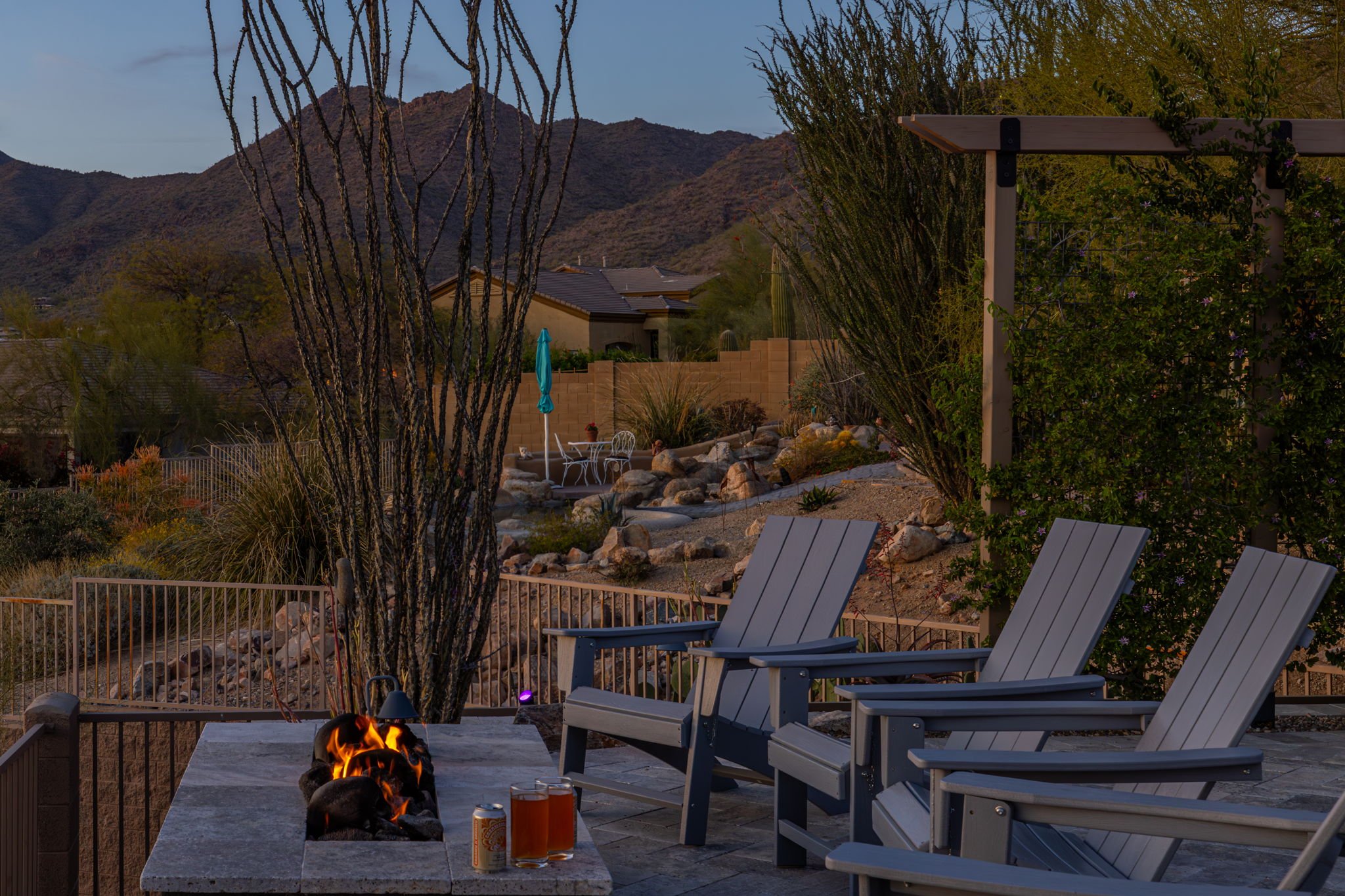 An outdoor patio at dusk with three gray Adirondack chairs, a fire pit with flames, two glasses of beer, a can, and various desert plants, trees, rocks, and a mountain landscape in the background.