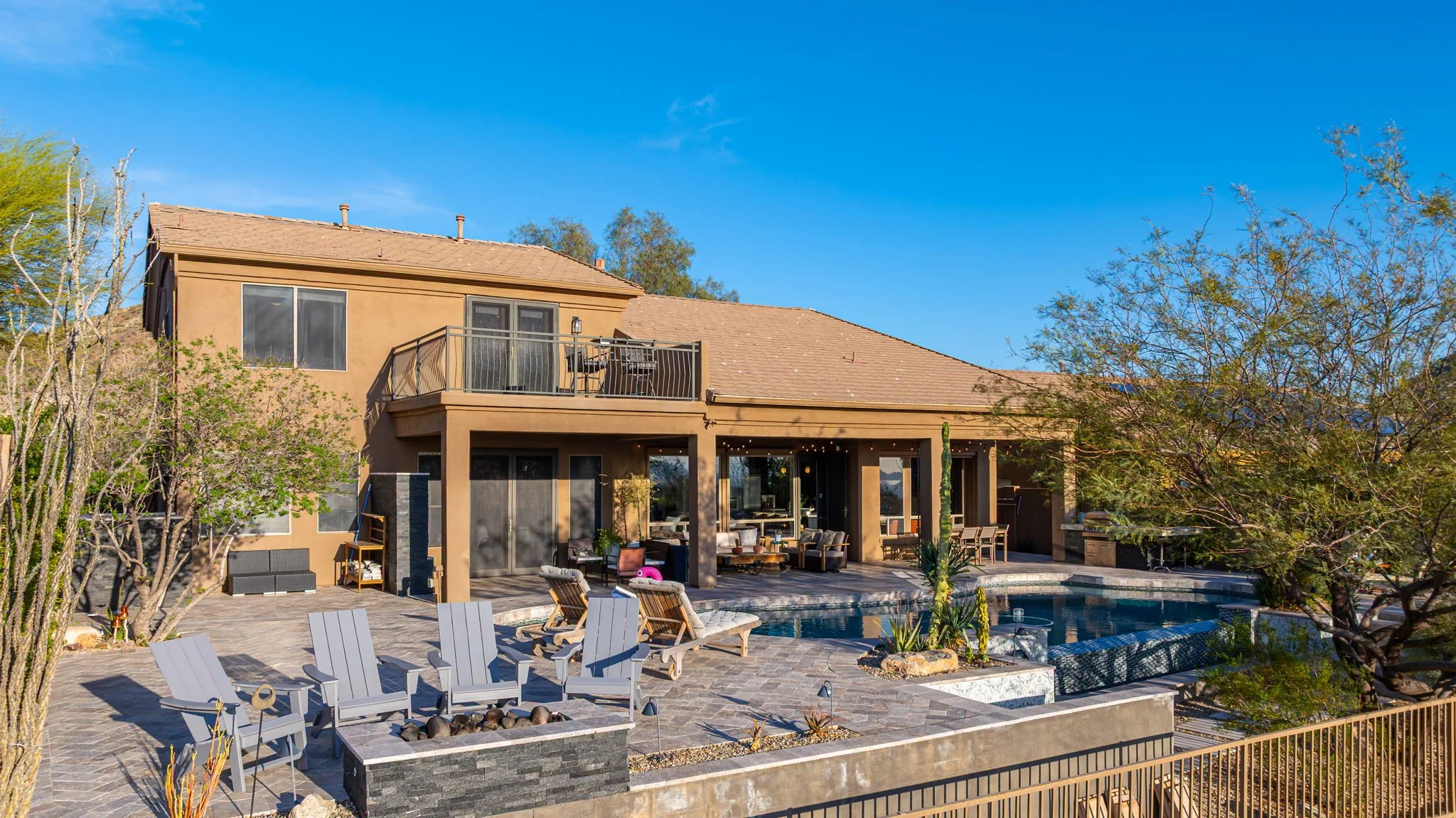 Backyard of a two-story house with a swimming pool, patio furniture, trees, and a clear blue sky.