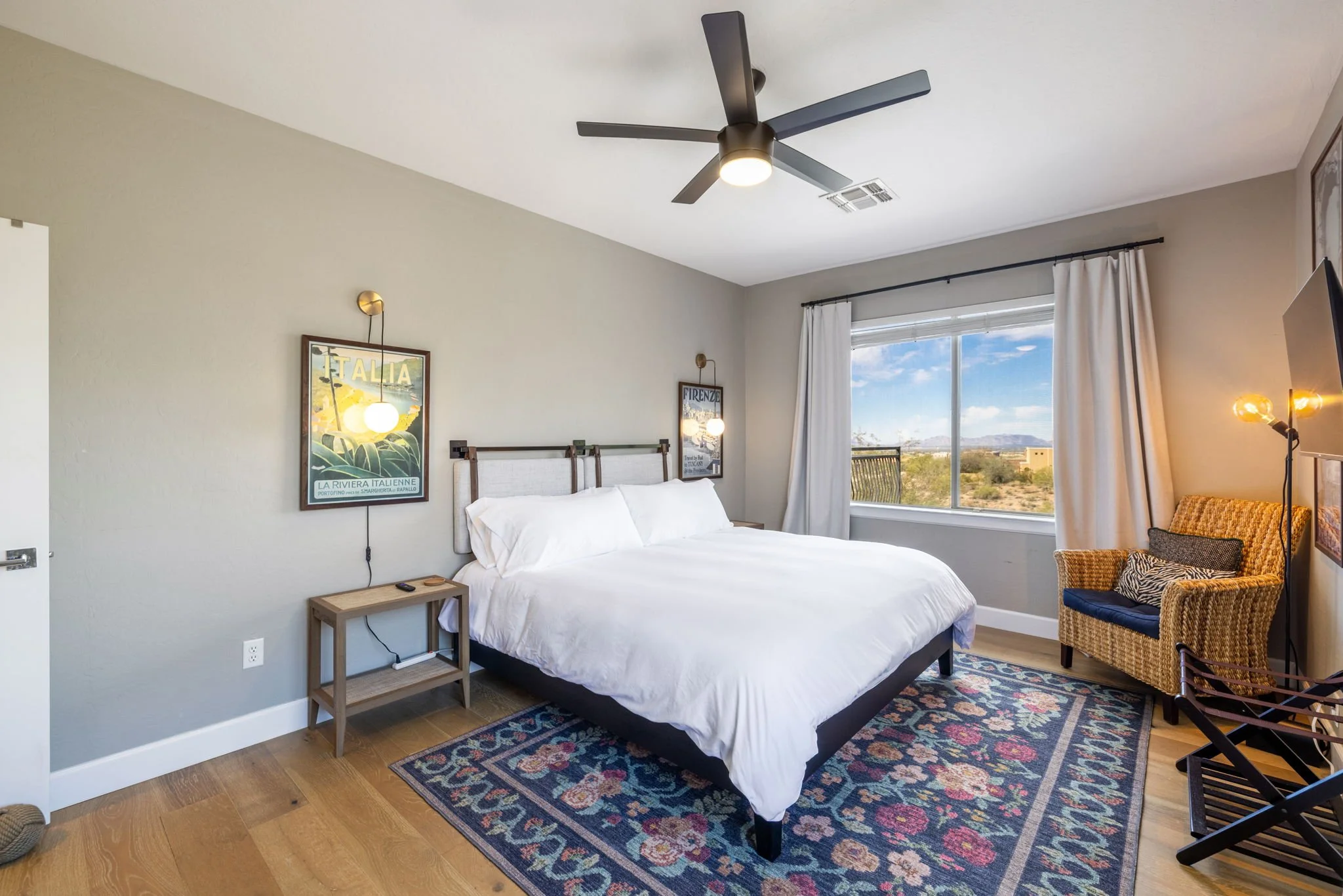 A modern bedroom with a large bed, white bedding, and a wooden headboard. There's a colorful area rug, a wicker armchair with a blue cushion, and a sliding glass window with white curtains showing a desert landscape outside. The room has a dark ceili