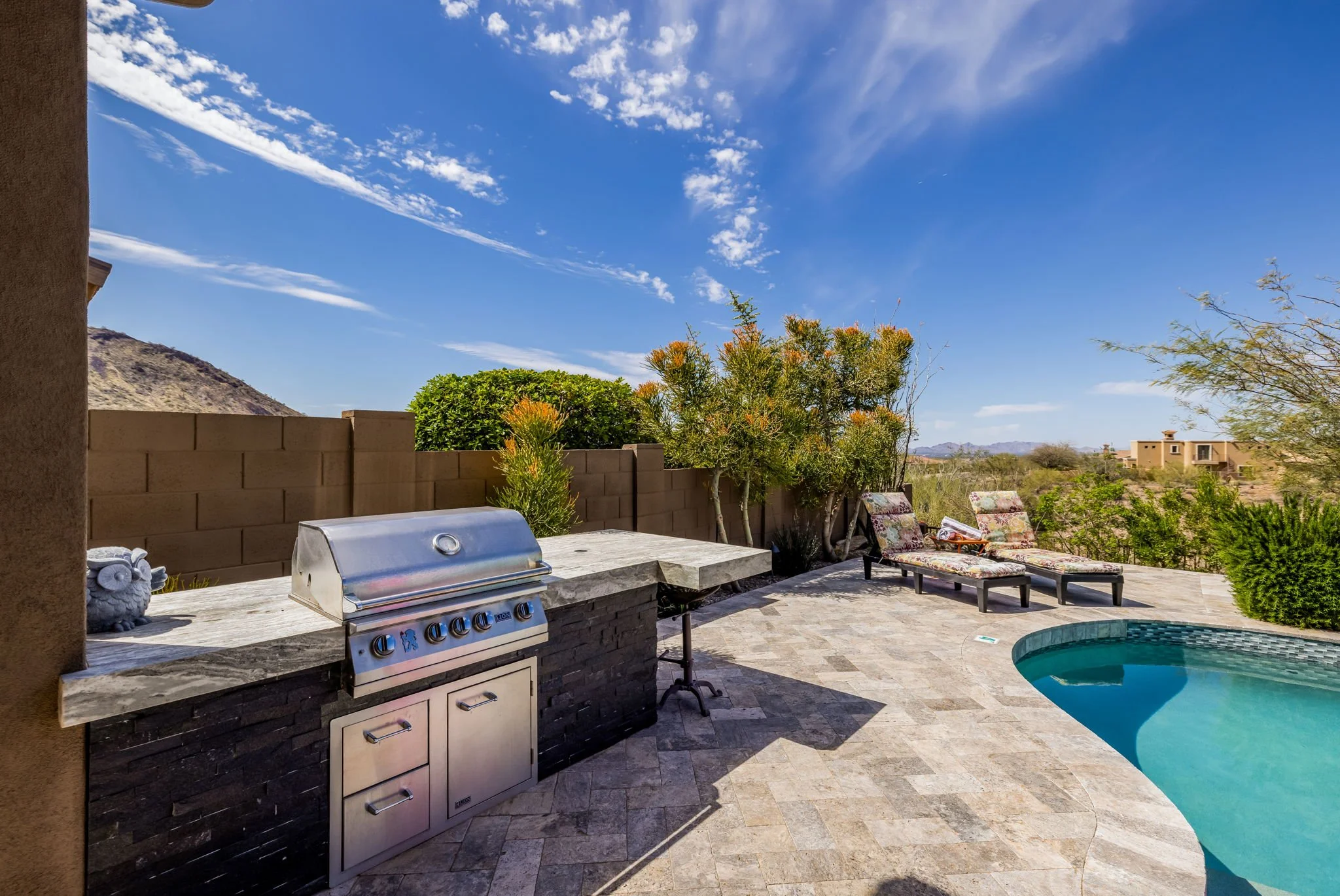 Outdoor backyard with pool, sun loungers, a built-in grill, and desert landscape in the background under a blue sky with scattered clouds.