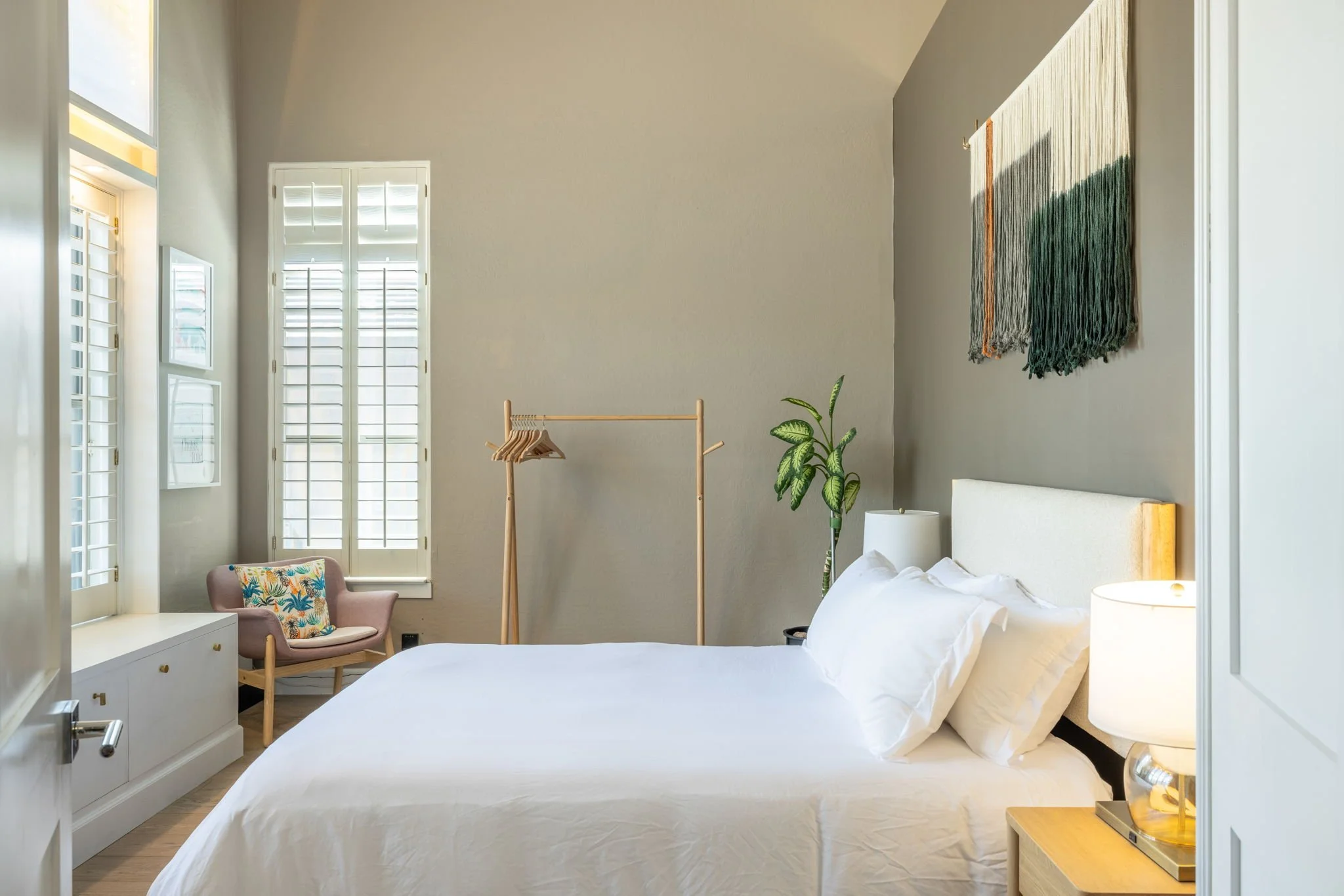 Bedroom with white bed, nightstand, lamp, window shutters, pink armchair with colorful pillow, indoor plant, hanging wall art, and wooden clothing rack.