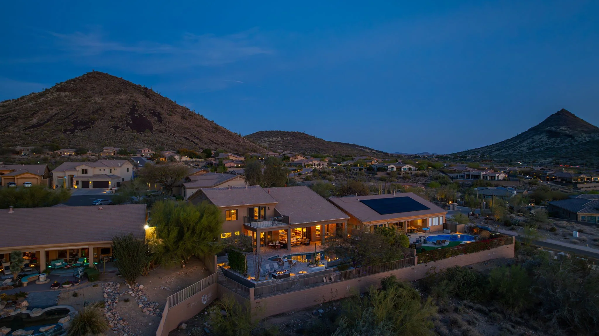 Aerial view of a residential neighborhood at dusk with mountains in the background, showcasing illuminated houses and outdoor spaces.