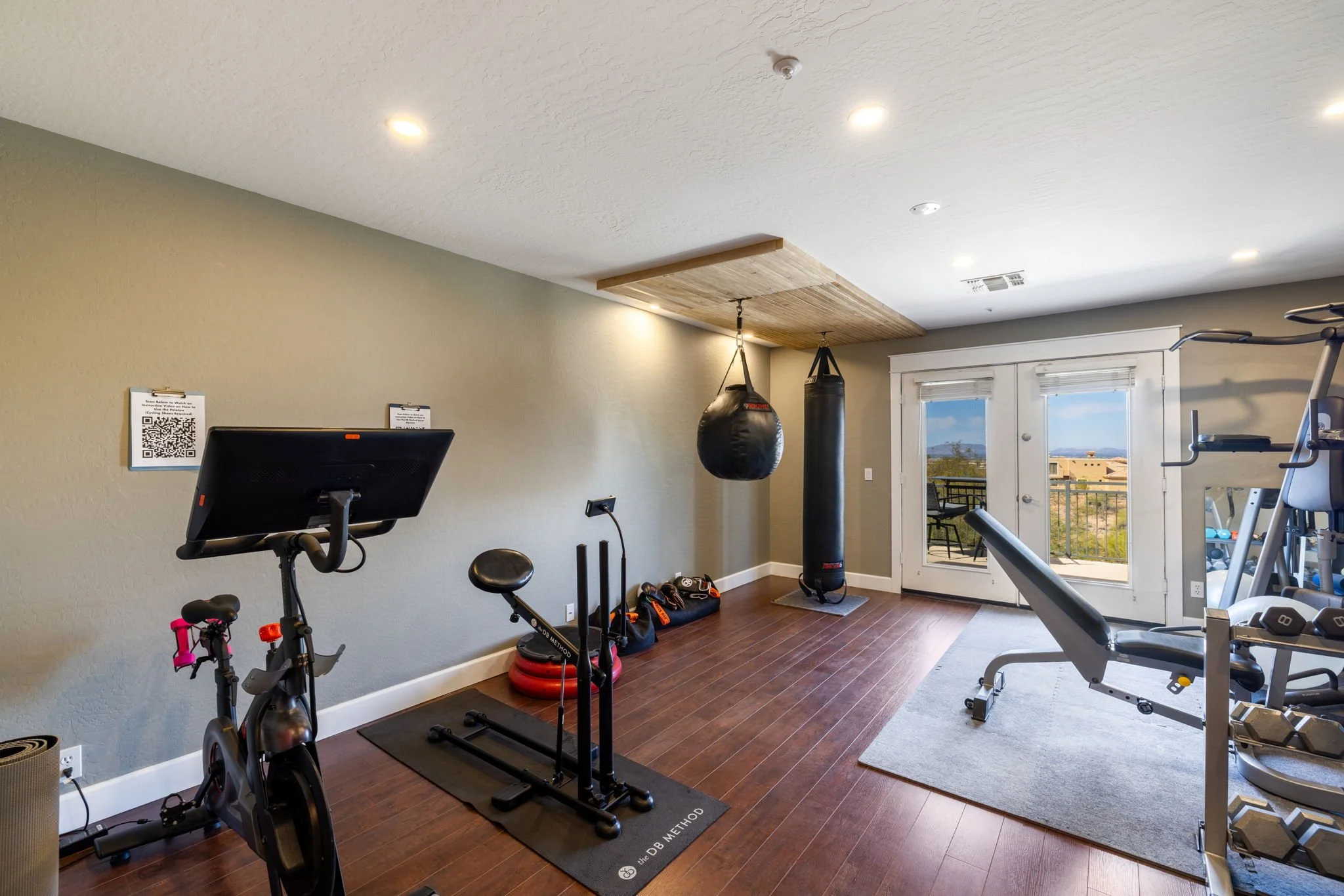 Home gym with exercise bike, punching bags, weight bench, and workout equipment, near glass doors leading to a balcony with outdoor scenery.