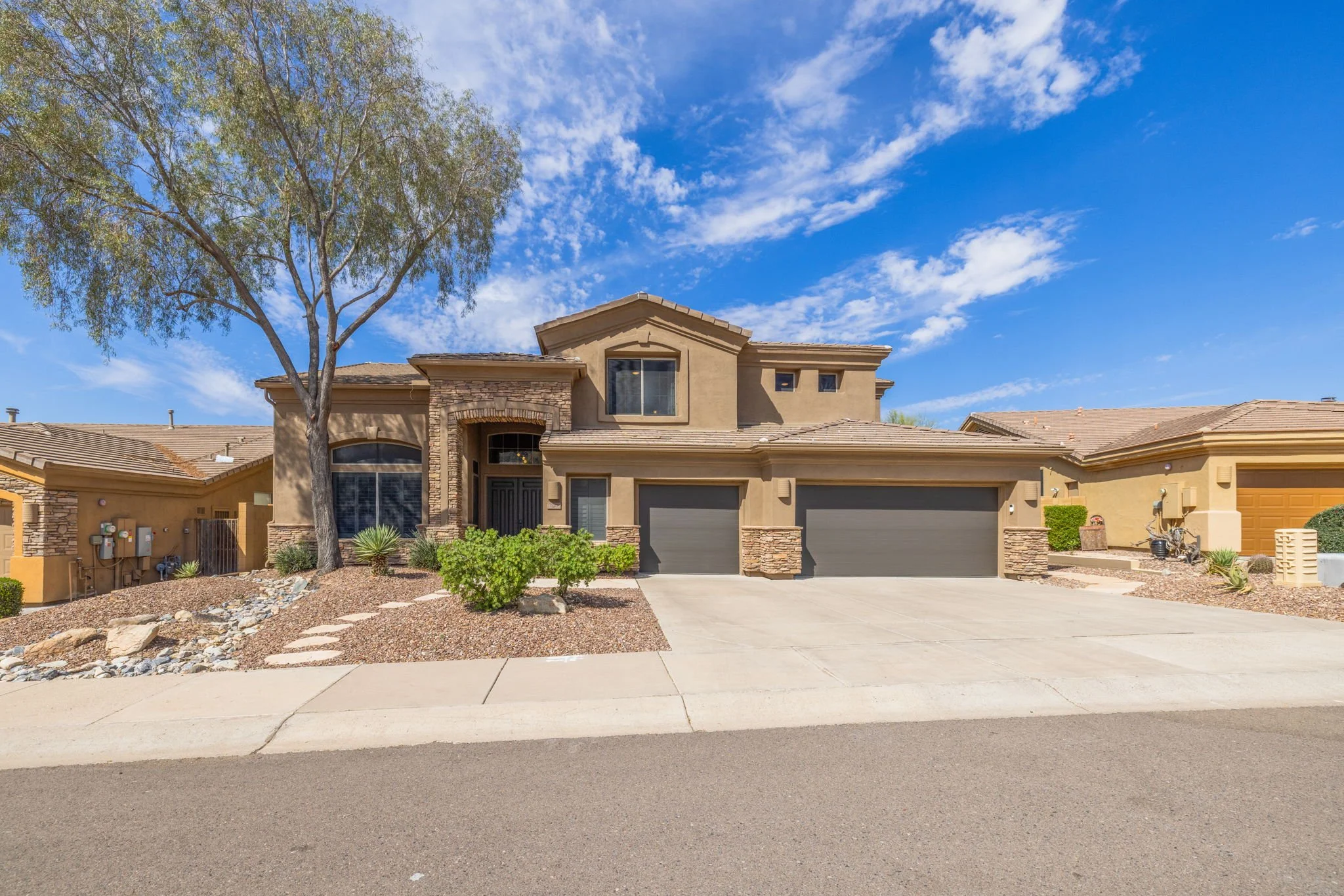 A modern two-story suburban house with a beige and brown exterior, stone accents, a three-car garage, and desert landscaping under a blue sky with scattered clouds.