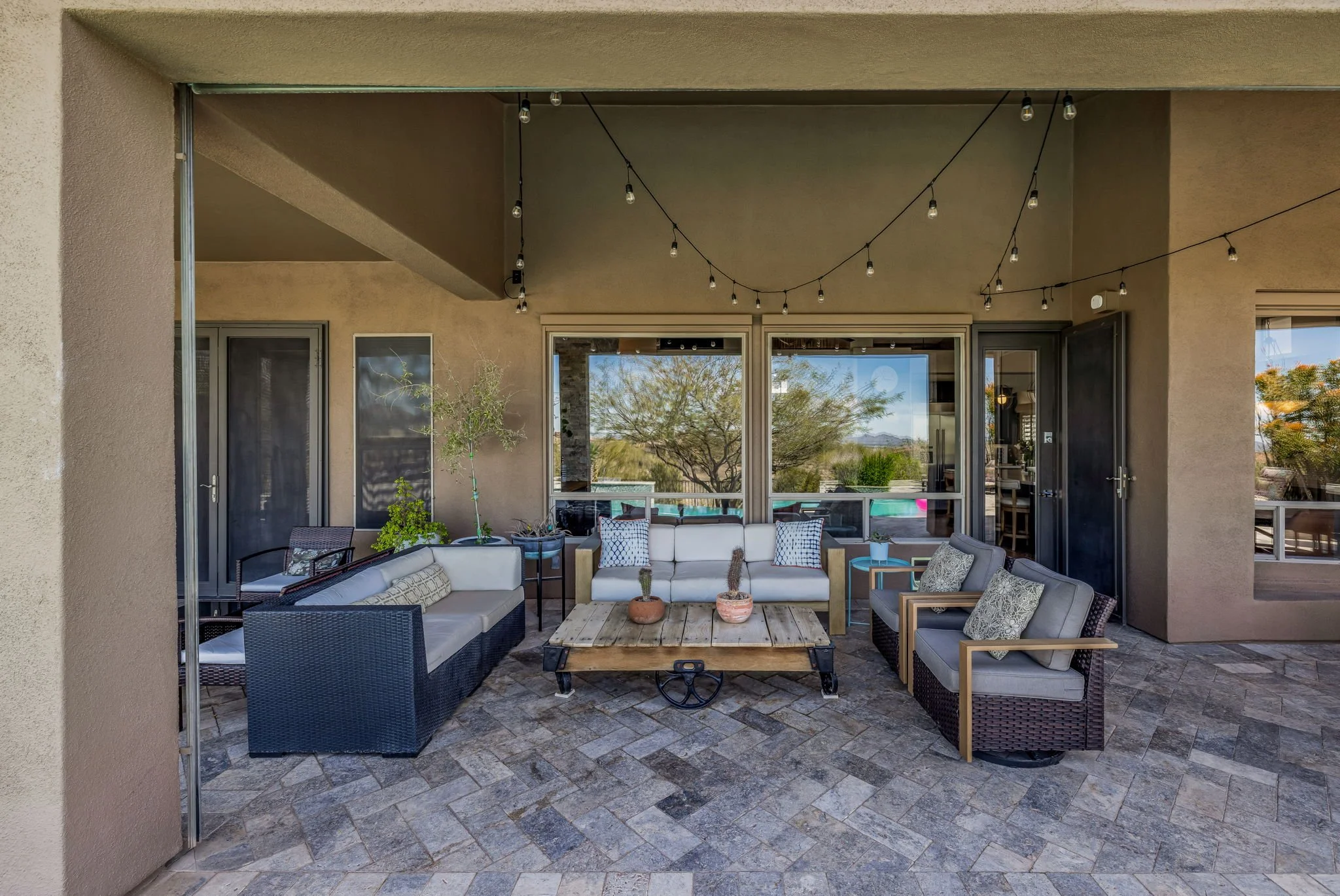 Covered outdoor patio with seating area, string lights, potted plants, and large windows reflecting trees.