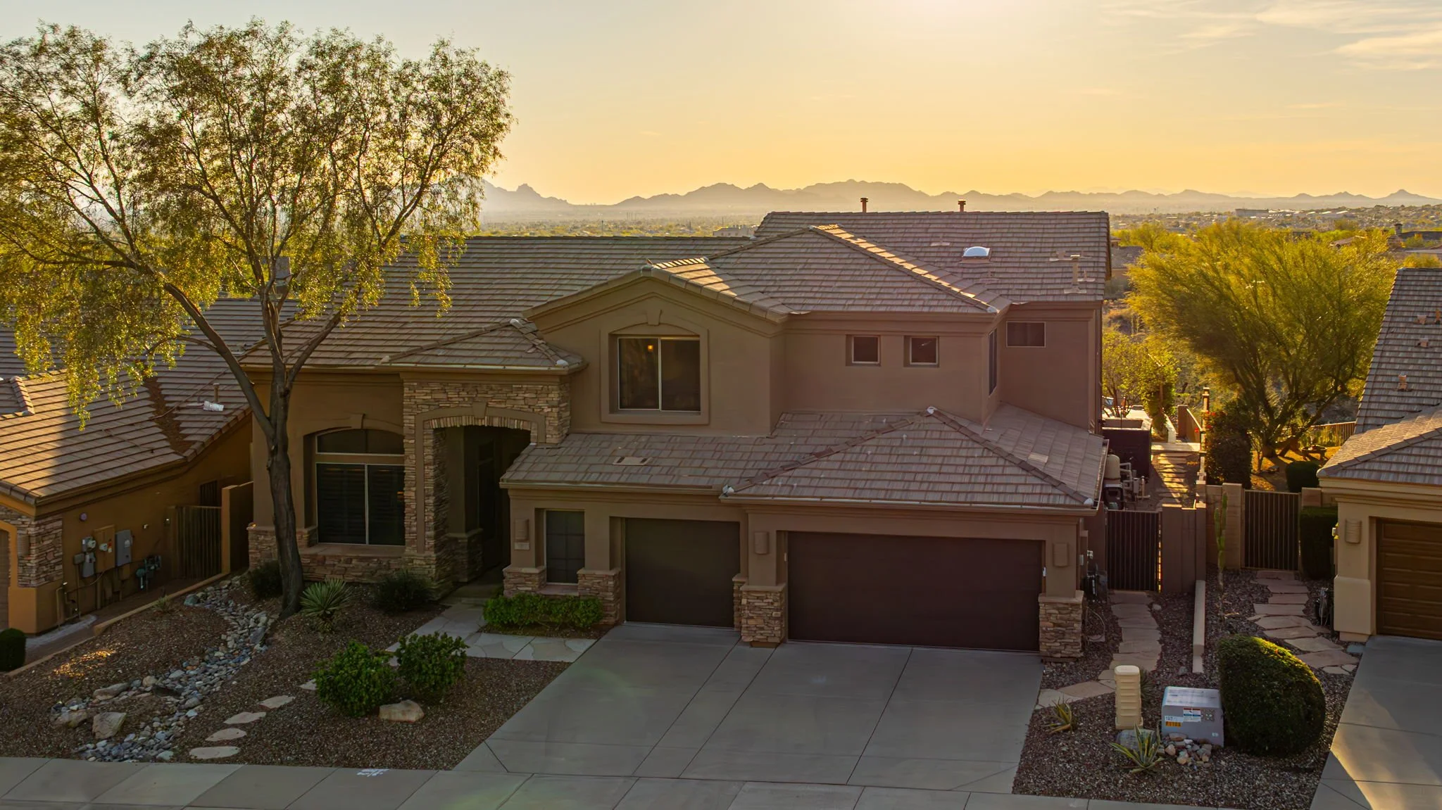 A two-story suburban house with a tile roof and stone accents, front yard with bushes and trees, driveway, and a sunset sky in the background.