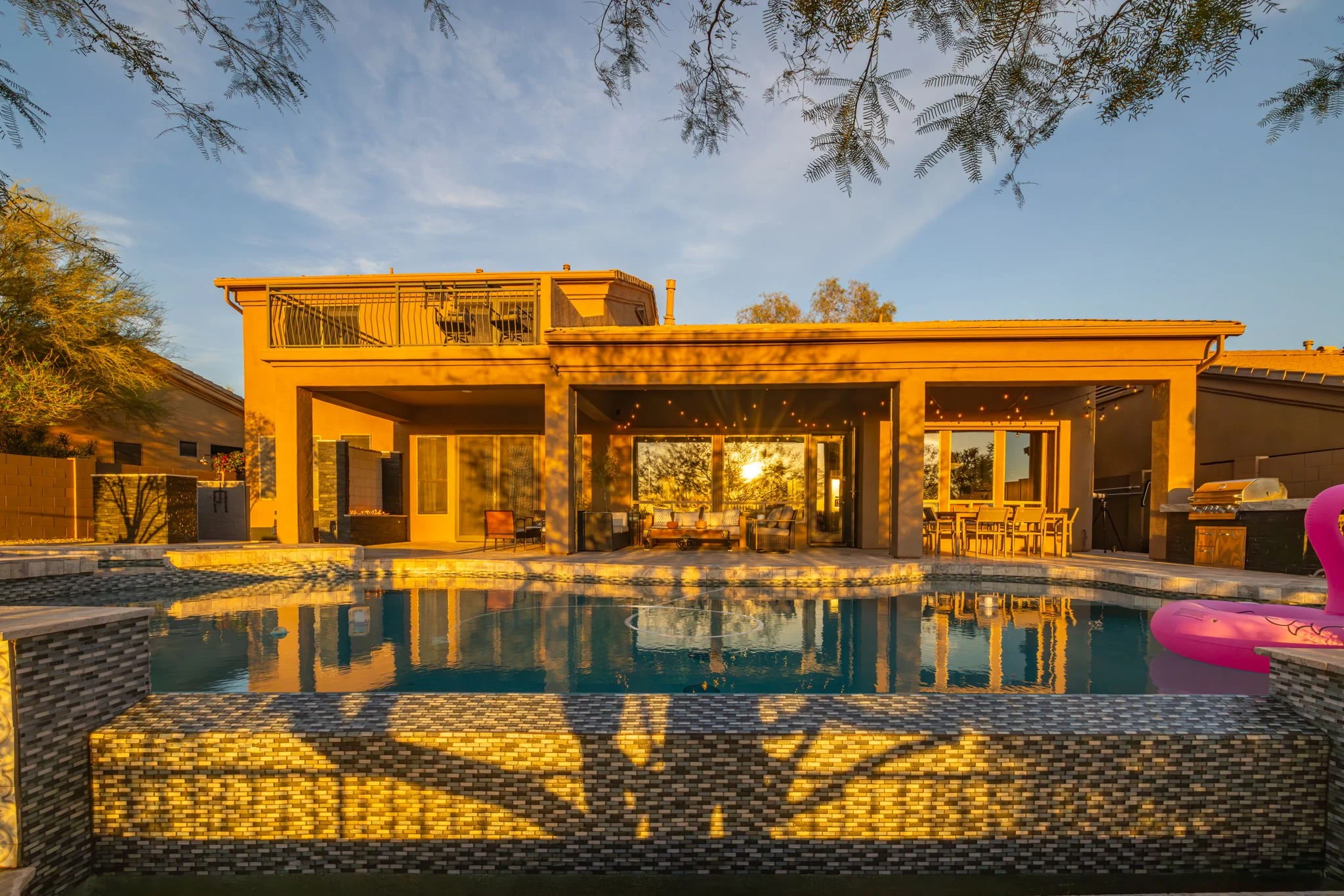 A modern house with a backyard swimming pool at sunset, surrounded by outdoor furniture and string lights, with a sunset reflected in the pool.