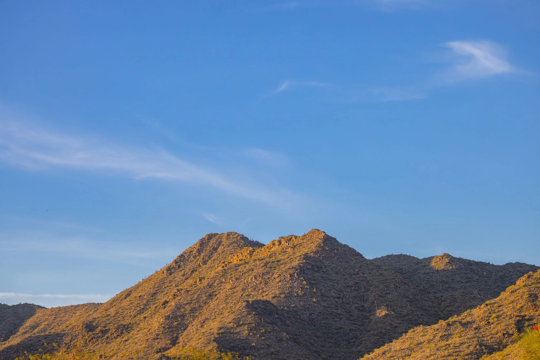 Sunlit desert mountain range under a blue sky with a few wispy clouds.