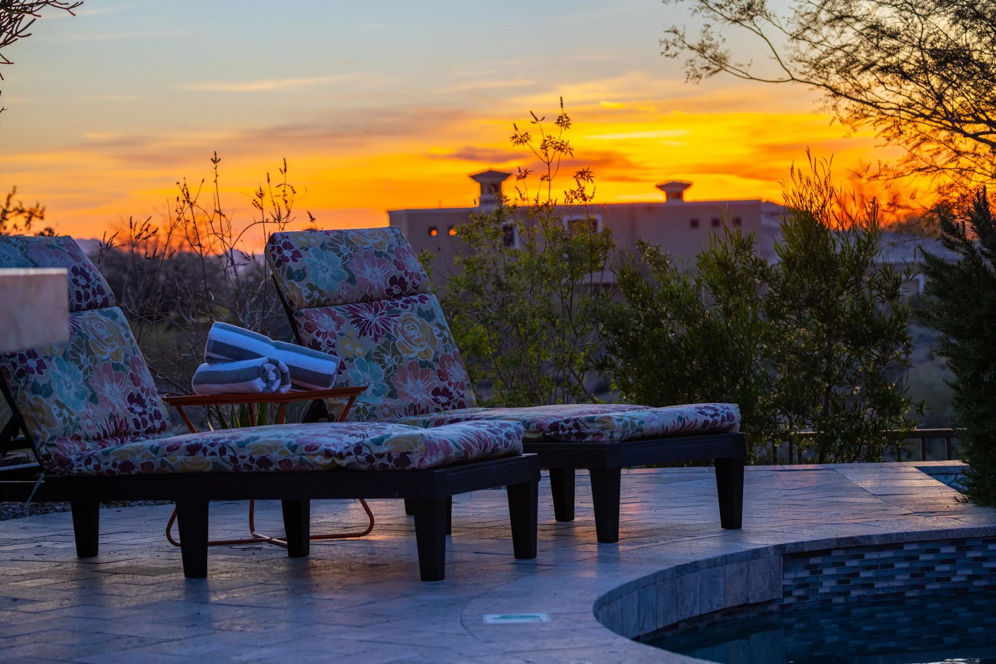 Outdoor poolside lounge area with floral cushioned chairs, a rolled towel, and a small table, overlooking a sunset with a colorful sky and trees in the background.