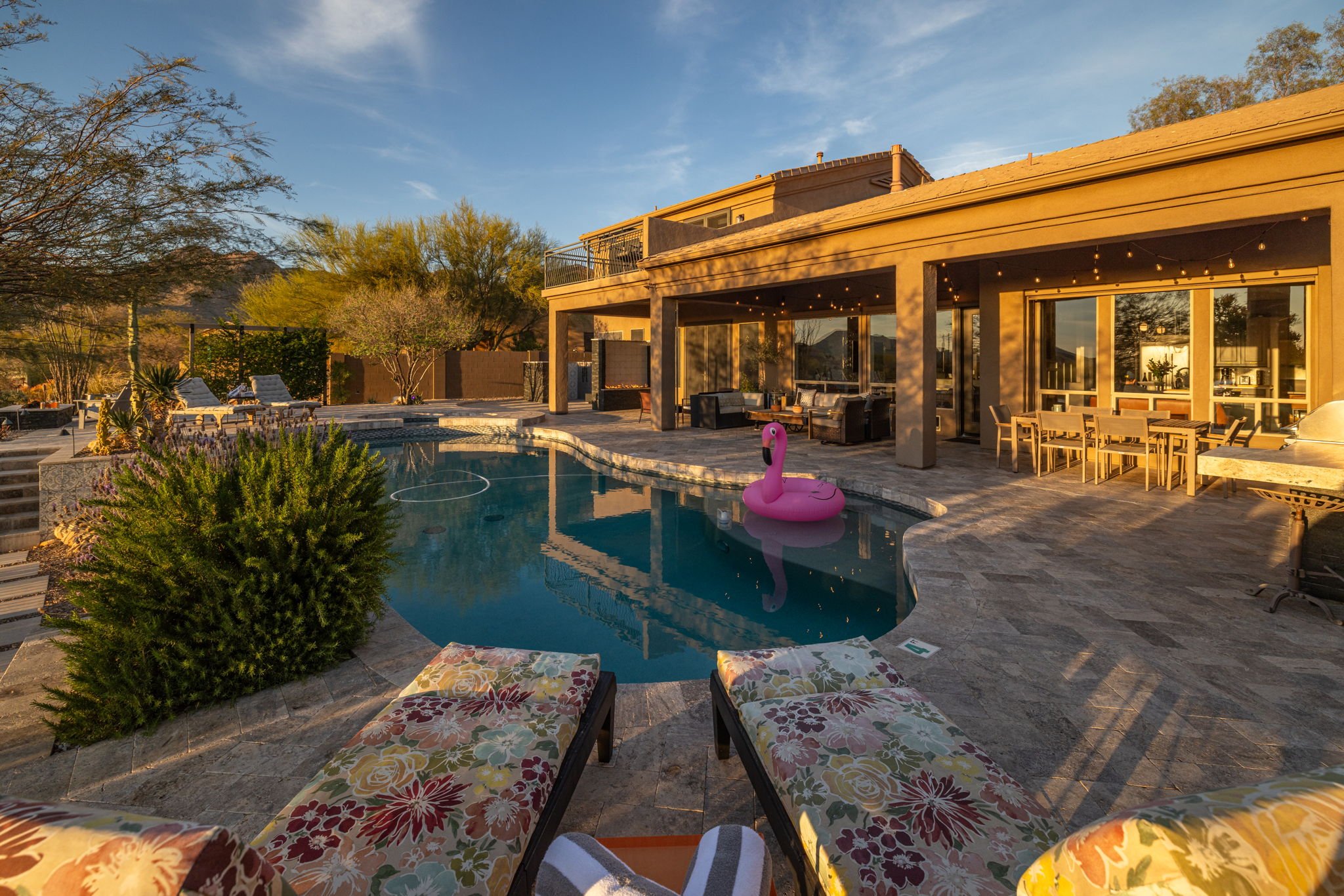 A backyard with a swimming pool, lounge chairs, and outdoor furniture in front of a beige house with large glass sliding doors, surrounded by trees and bushes, under a clear sky in warm sunlight.