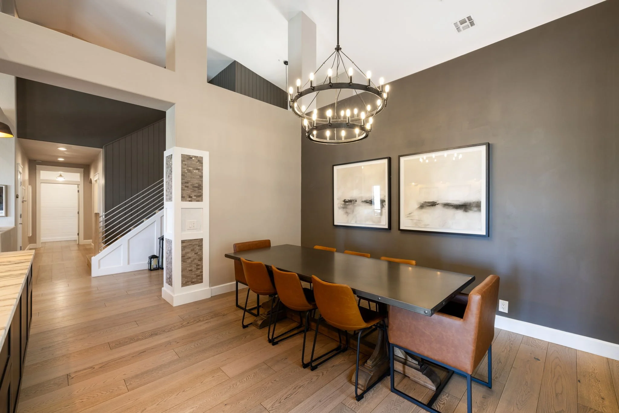 Modern dining room with a large rectangular table, six orange chairs, and one brown leather chair, two abstract black and white paintings on a gray wall, a black chandelier with multiple bulbs hanging above, hardwood floors, and a staircase with whit