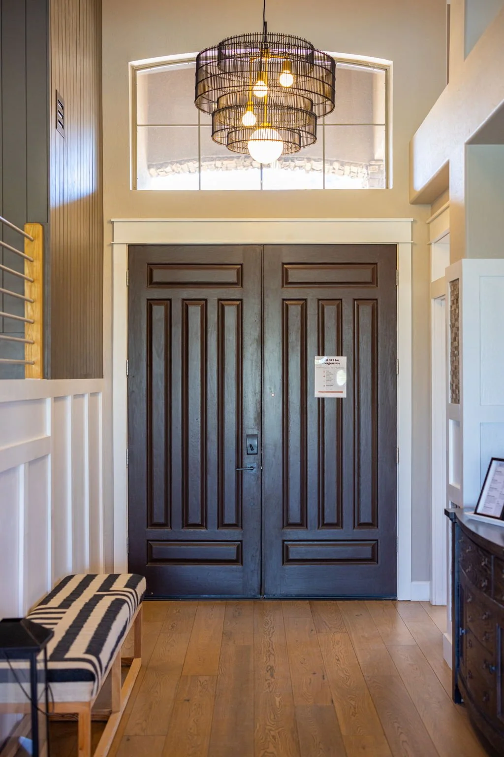 A doorway with a large set of double wooden doors. Above the doors is a window with a modern chandelier hanging in front of it. To the left is a light-colored wall with a wooden staircase rail and a bench with a black-and-white striped cushion. To th