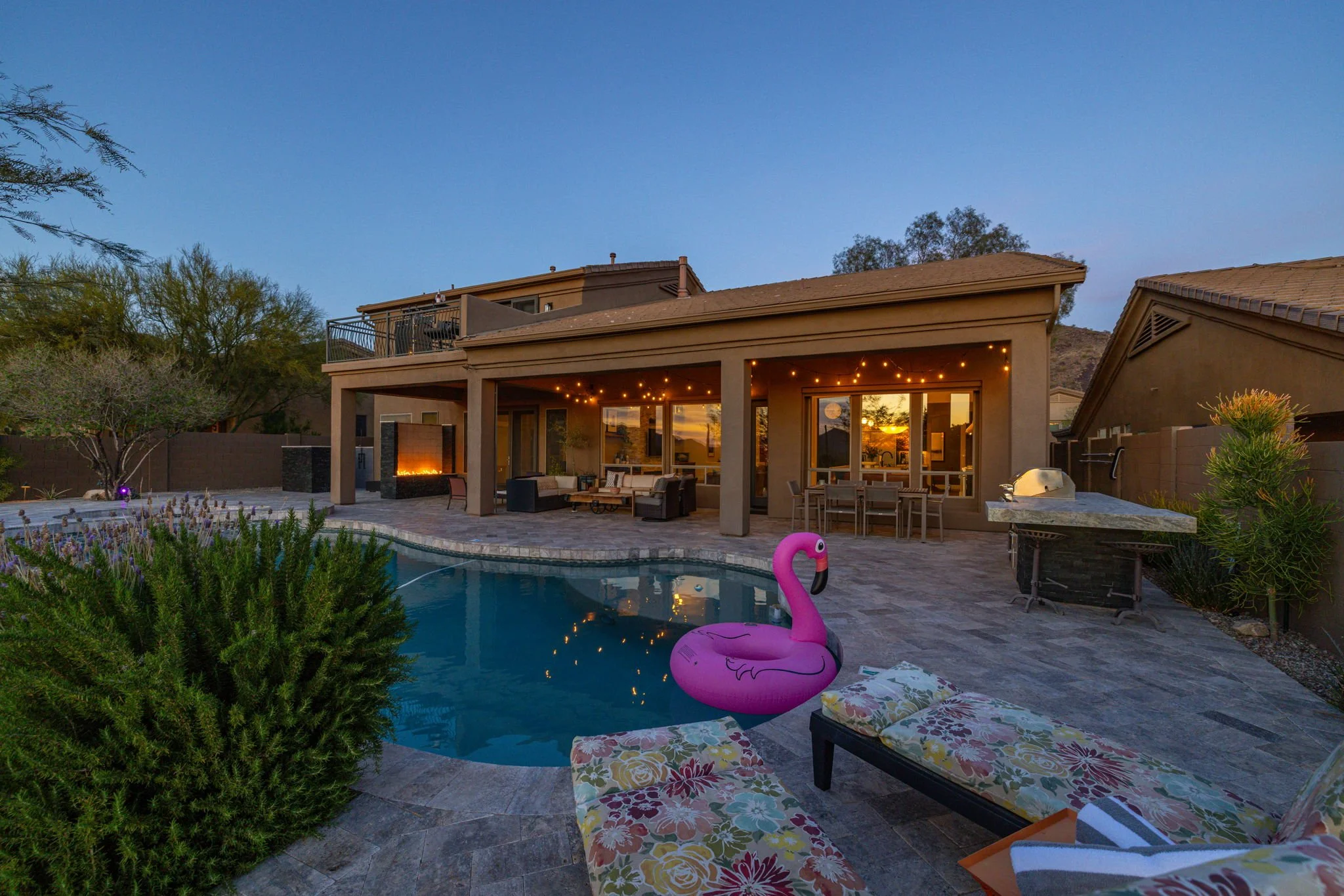 Backyard with swimming pool, inflatable pink flamingo, lounge chairs, patio furniture, outdoor fireplace, grill, and house with string lights at dusk.