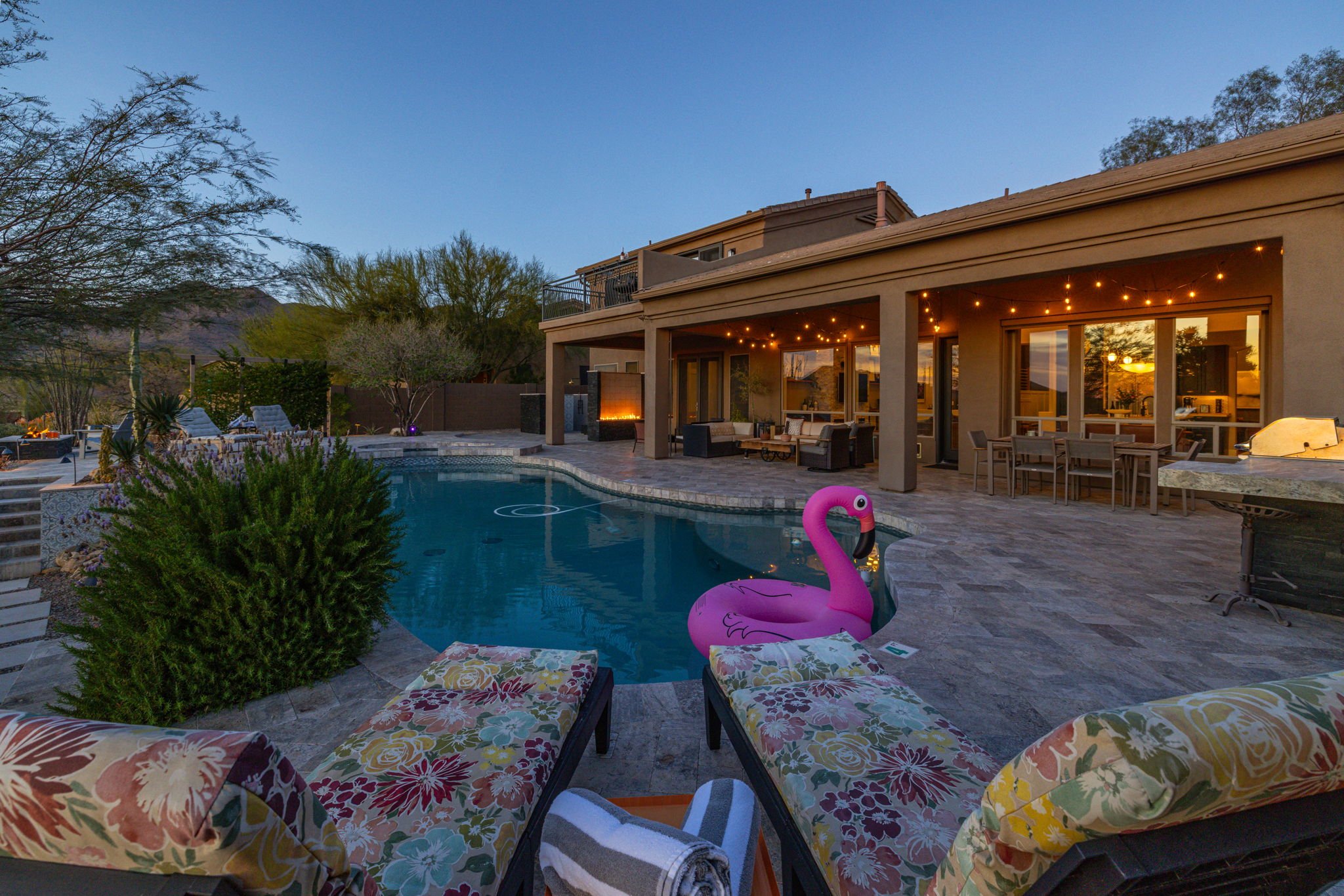 Backyard with a swimming pool, lounge chairs with floral cushions, a pink flamingo float, patio furniture, string lights, trees, and a house with large windows at dusk.
