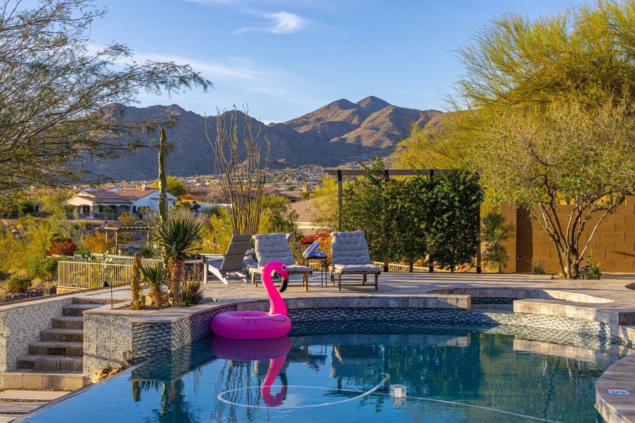 An outdoor backyard pool area with lounge chairs, a pink flamingo-shaped inflatable float, and desert mountain scenery in the background during daytime.
