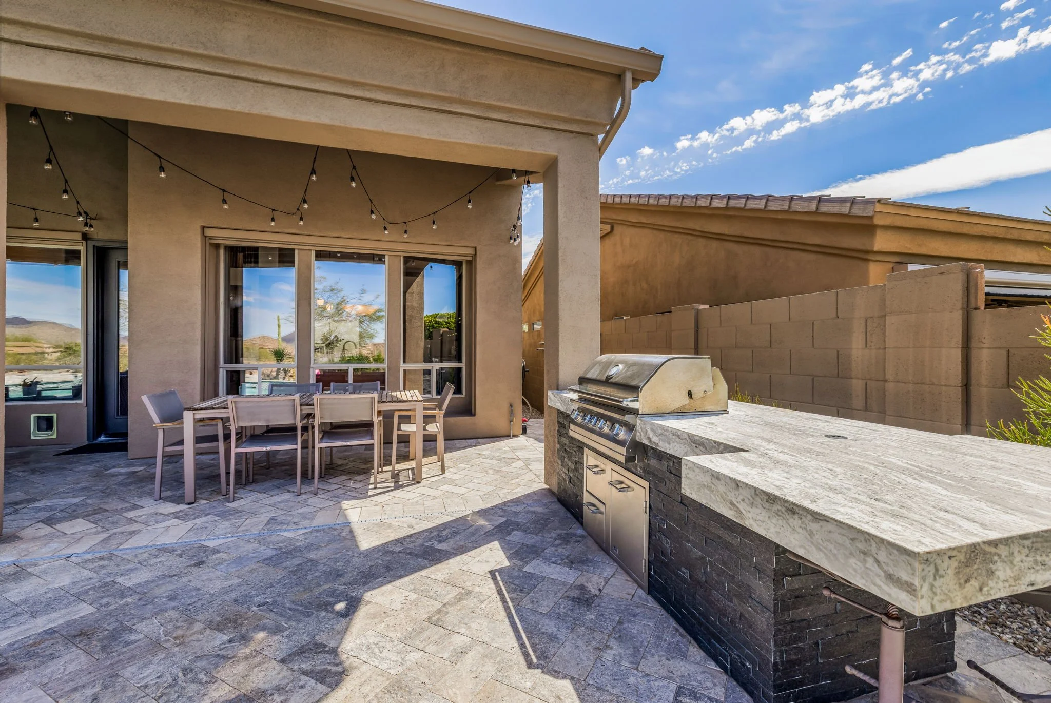 Outdoor patio with dining table, string lights, and built-in grill under a covered area, with a view of neighboring houses and desert landscape in the background.