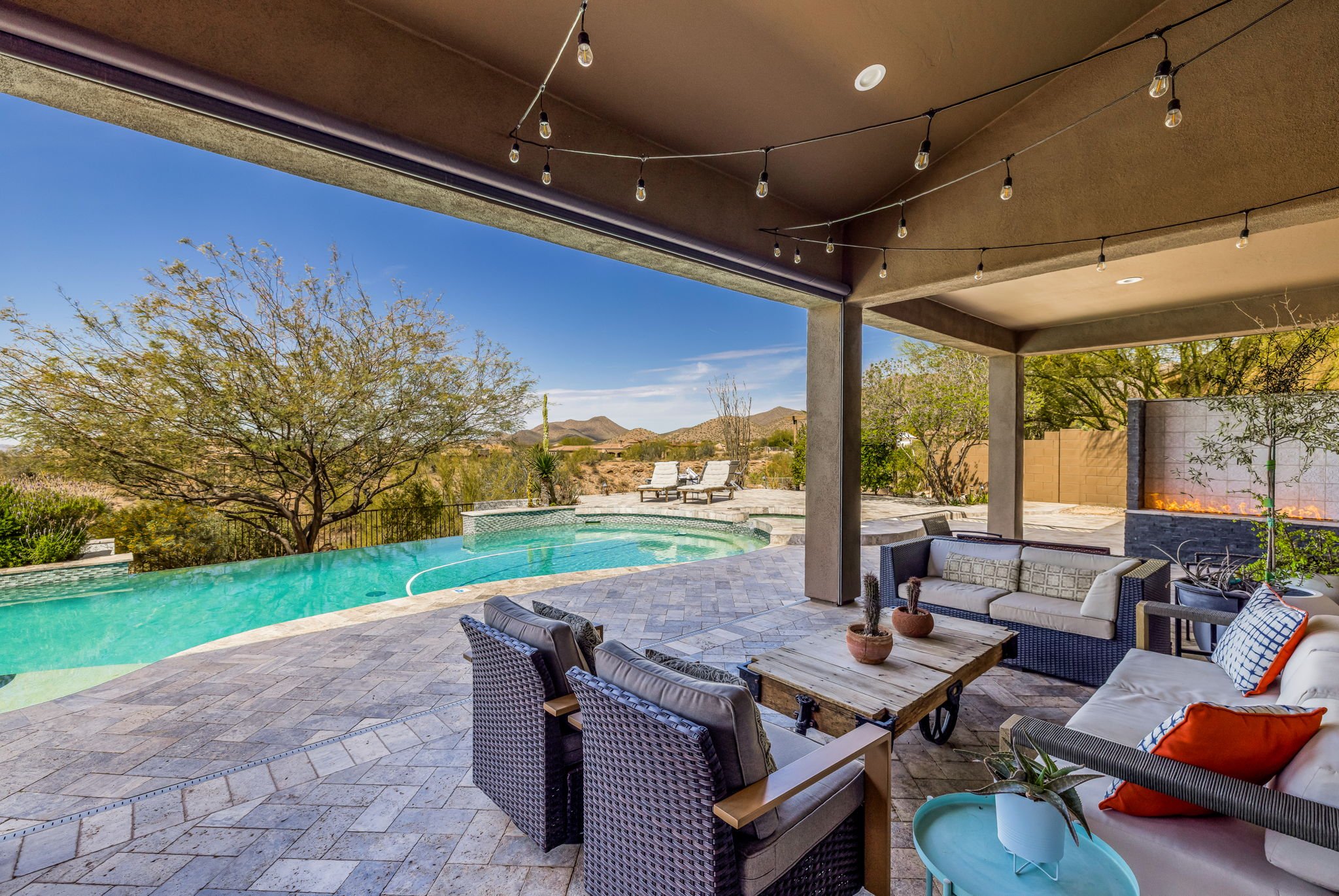 Covered patio with outdoor seating, string lights, and a view of a pool and desert landscape with mountains and trees.