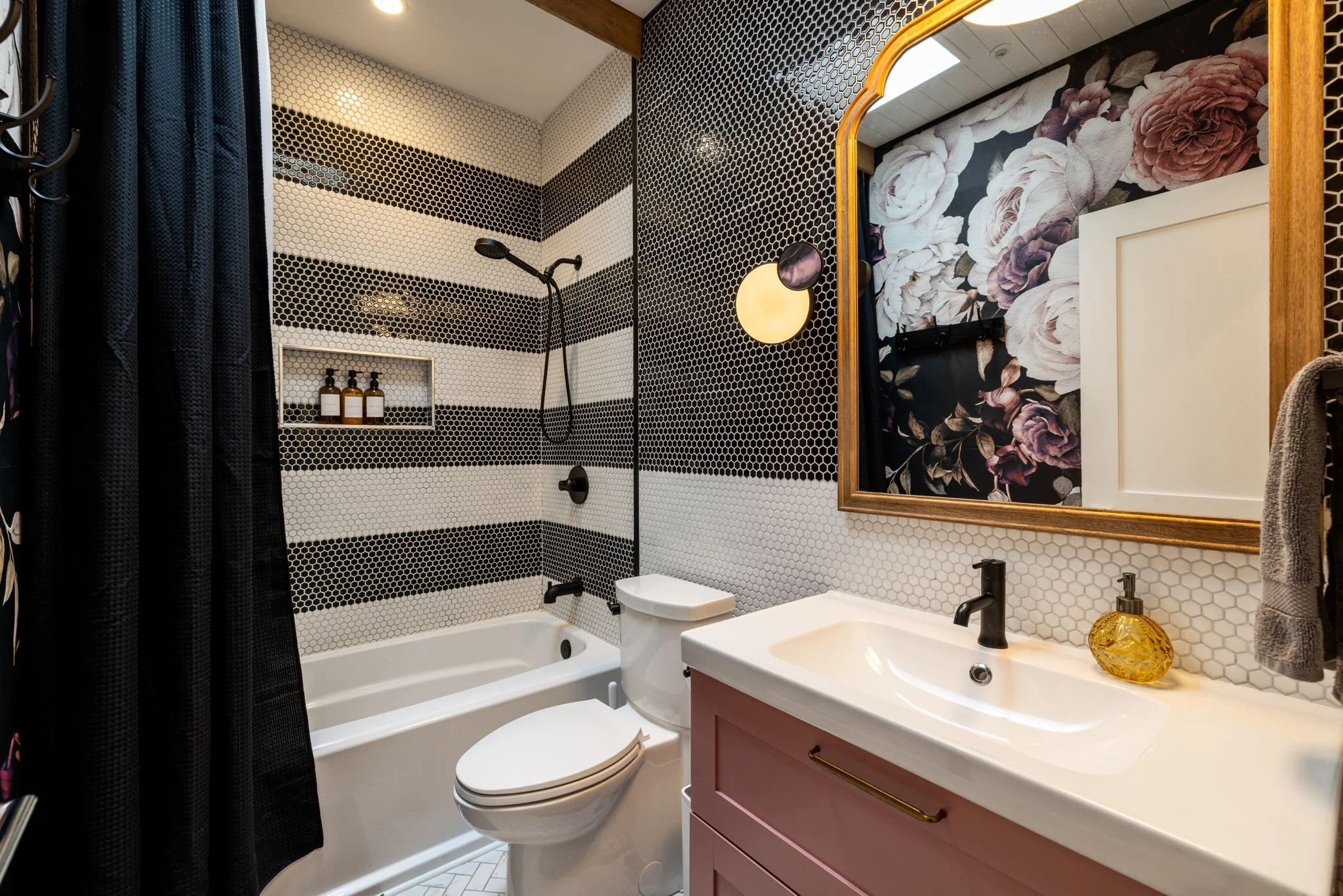 Bathroom with black and white striped hexagon tile walls, a bathtub with a black showerhead, a toilet, a pink vanity with a white sink, a large mirror with a floral pattern, a yellow decorative soap dispenser, and a dark gray hand towel.