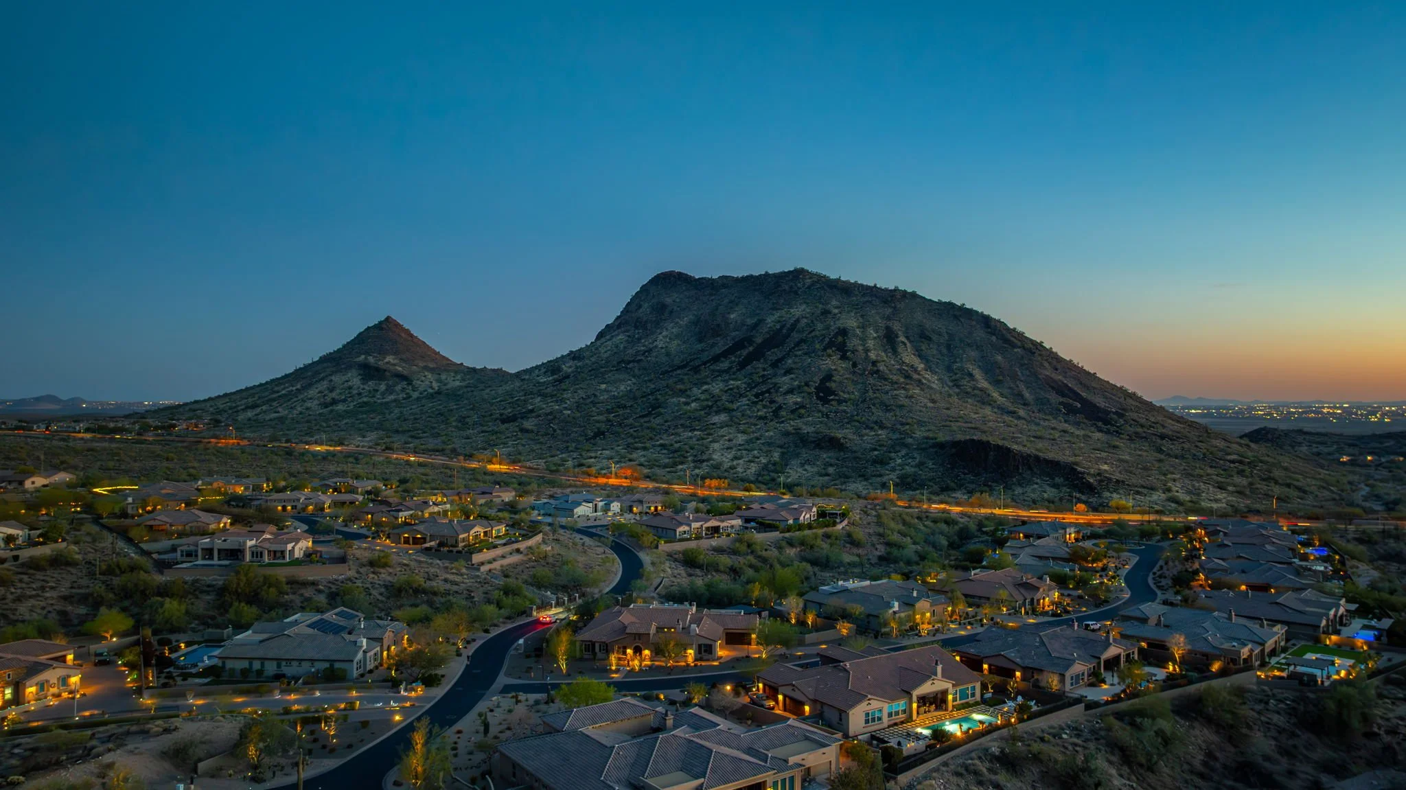 Twilight view of a suburban neighborhood with houses and streetlights in front of a mountain landscape with two prominent peaks.