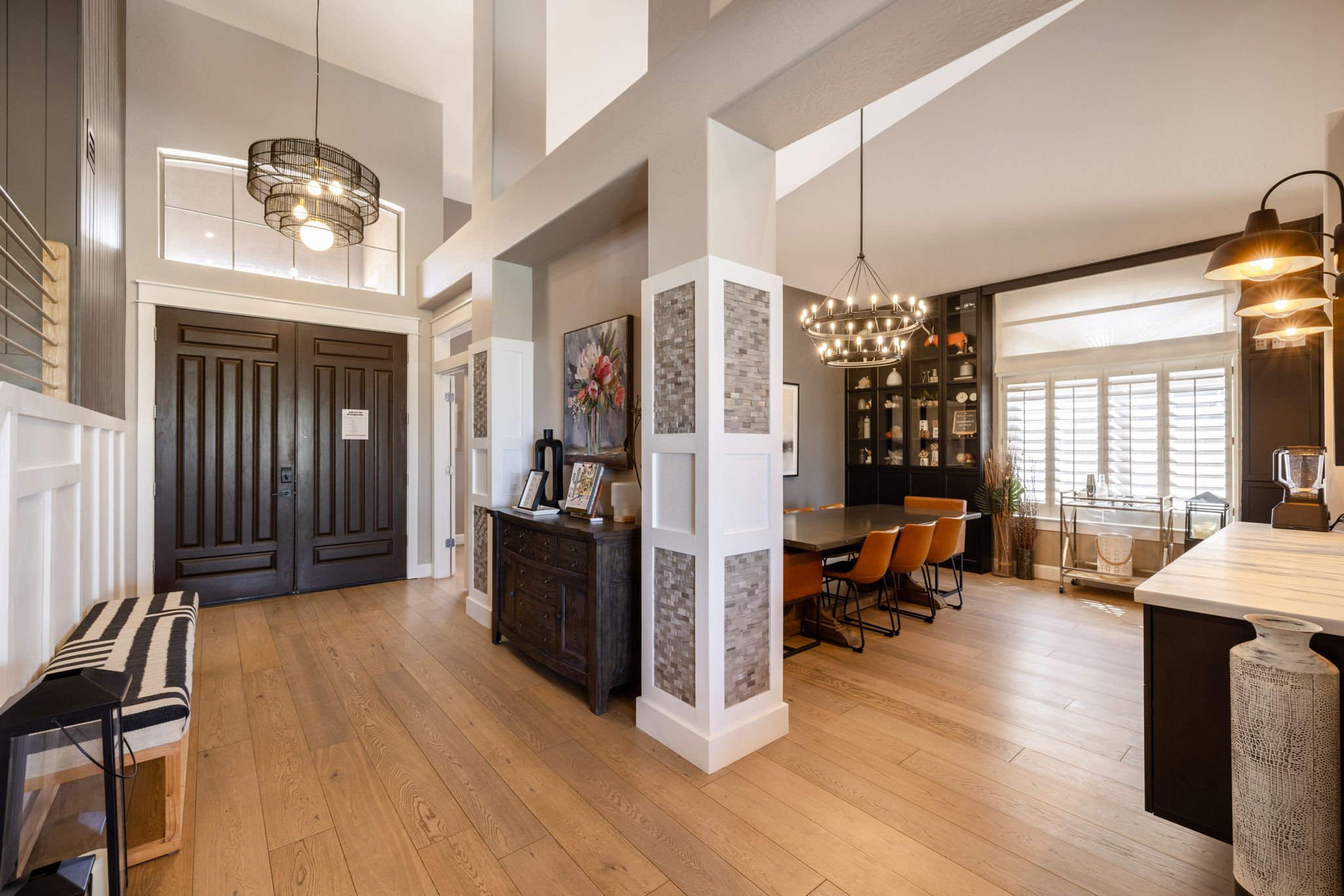 The photo shows a modern dining area with a black cabinet displaying pottery and decorative items, a large dining table with orange chairs, a window with white shutters, and contemporary lighting fixtures, including a chandelier and wall lights. The 