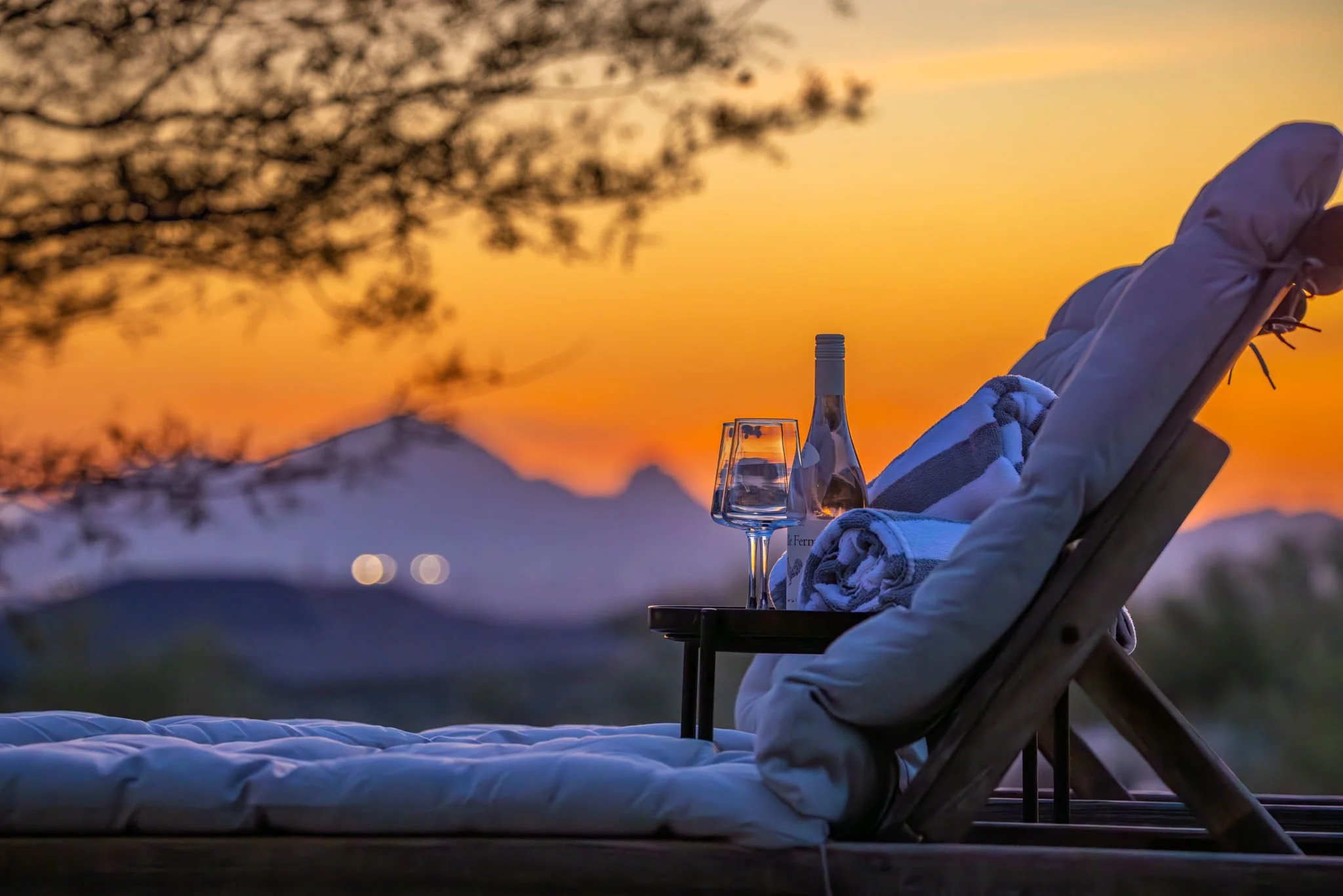 A lounge chair with a rolled towel, a water bottle, and a glass on a small table, set outdoors at sunset with mountain silhouettes in the background.