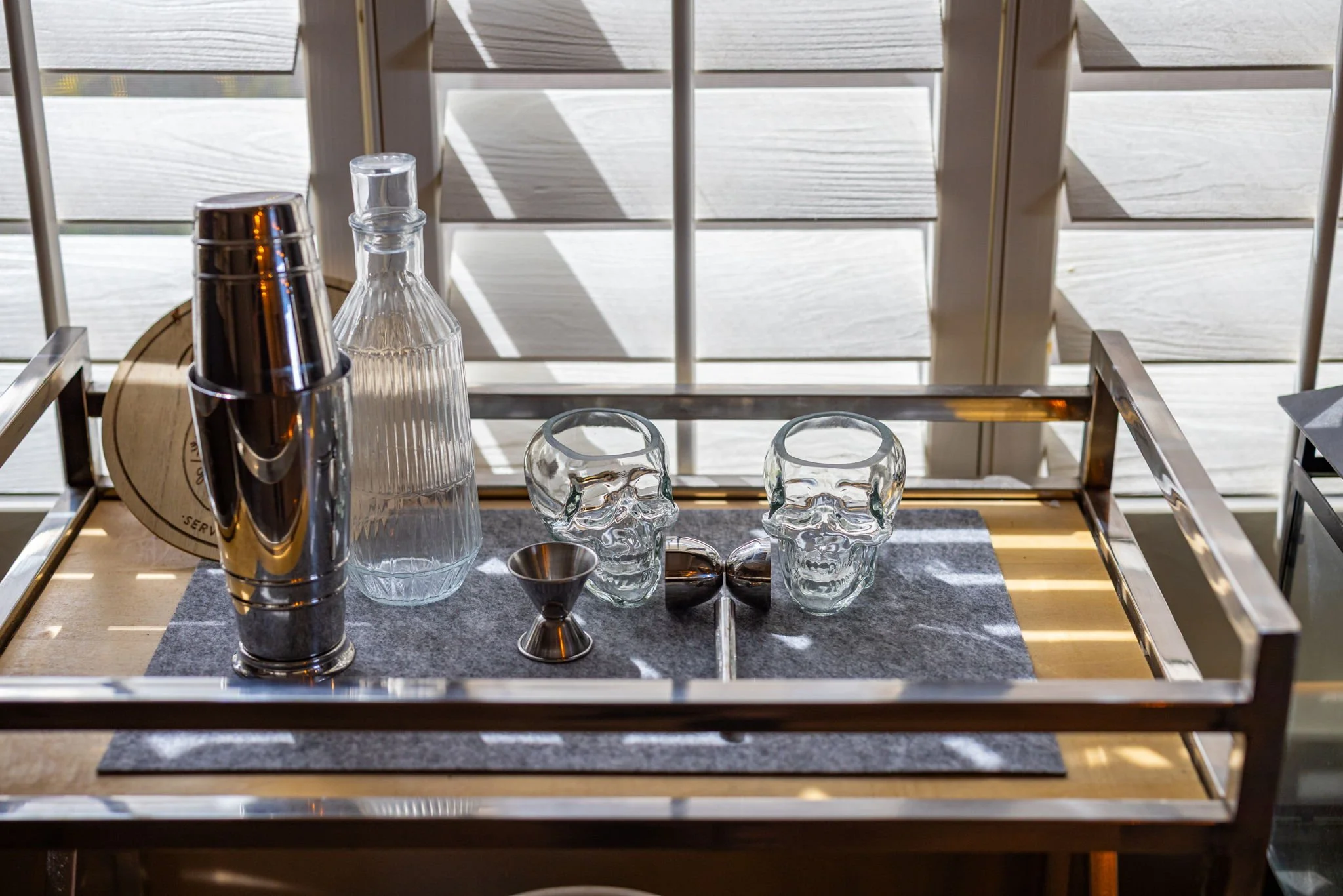 A bar cart with a silver shaker, glass decanter, two glass skulls, a small metal jigger, and a pair of metal ice tongs, set on a dark mat, with sunlight and shadows on a white wooden background.