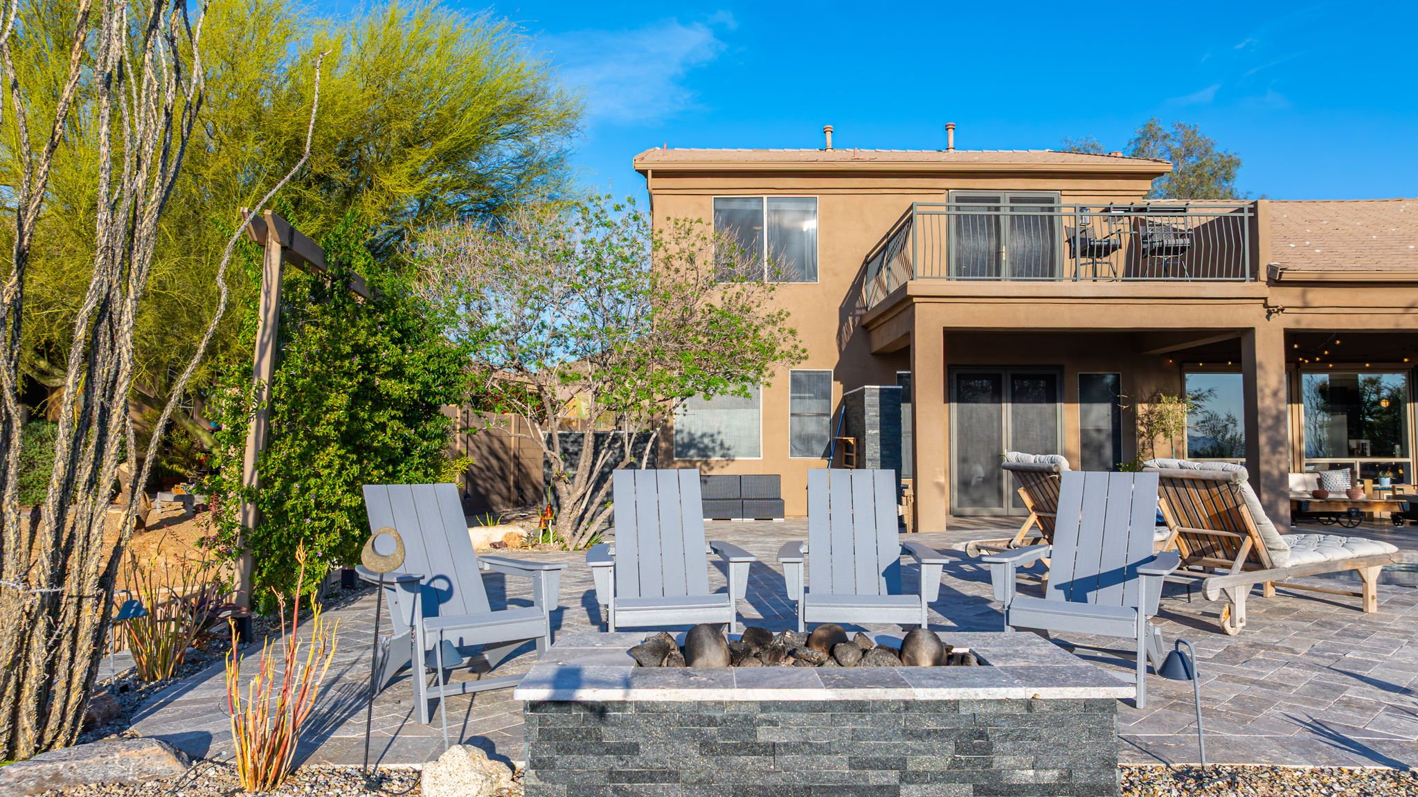 Backyard patio with gray Adirondack chairs around a fire pit, lounge chairs, shrubs, trees, a two-story house with a balcony, and clear blue sky.