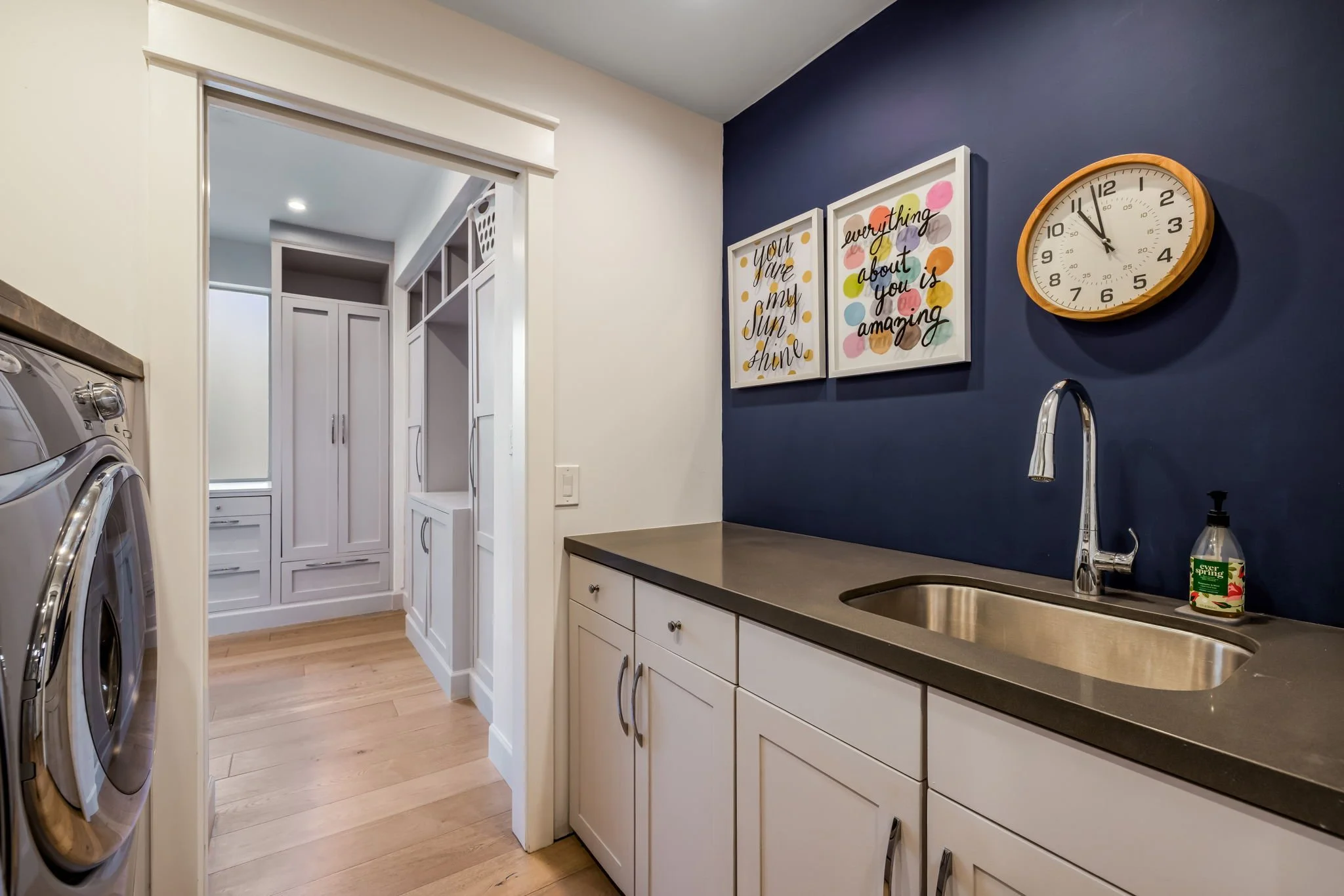 Laundry room with a white cabinet, a gray countertop, a stainless steel sink, a soap dispenser, two framed colorful quote posters on a navy blue wall, a round wall clock, and a doorway leading to a storage closet with built-in shelves.