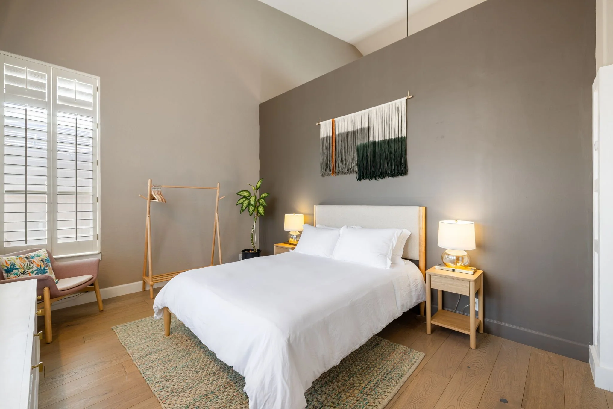 A bedroom with a white bed, two bedside tables with lamps, a hanging wall art, a chair, a potted plant, a window with white shutters, and a wooden clothes rack.