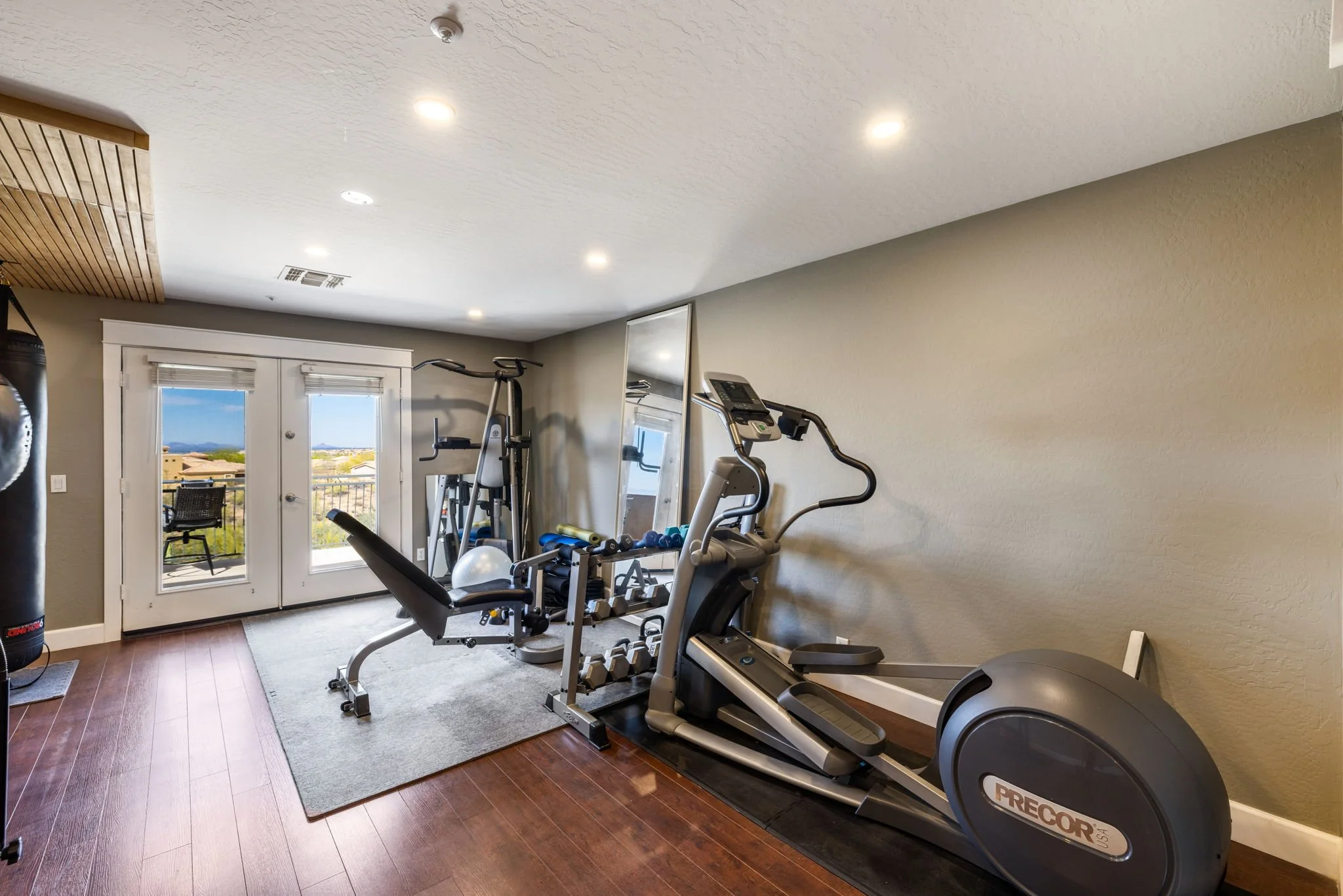 Home gym with treadmill, elliptical, weight bench, dumbbells, and a large mirror near glass double doors leading to a balcony.