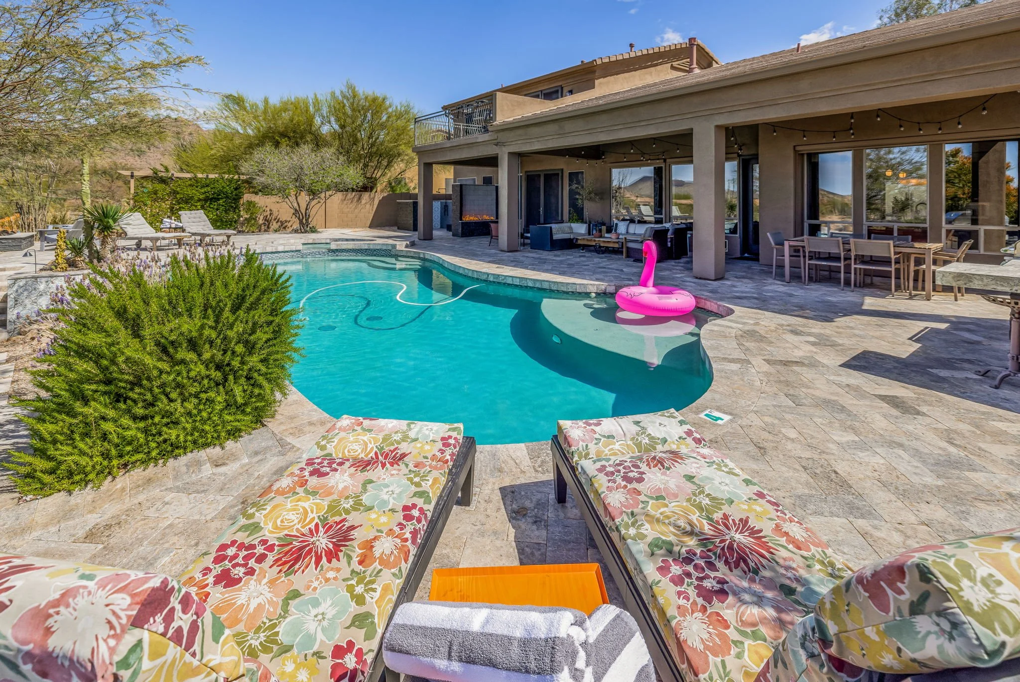 Backyard with a house featuring a covered patio, a swimming pool with a pink flamingo float, lounge chairs with floral cushions, and various outdoor furniture and plants.