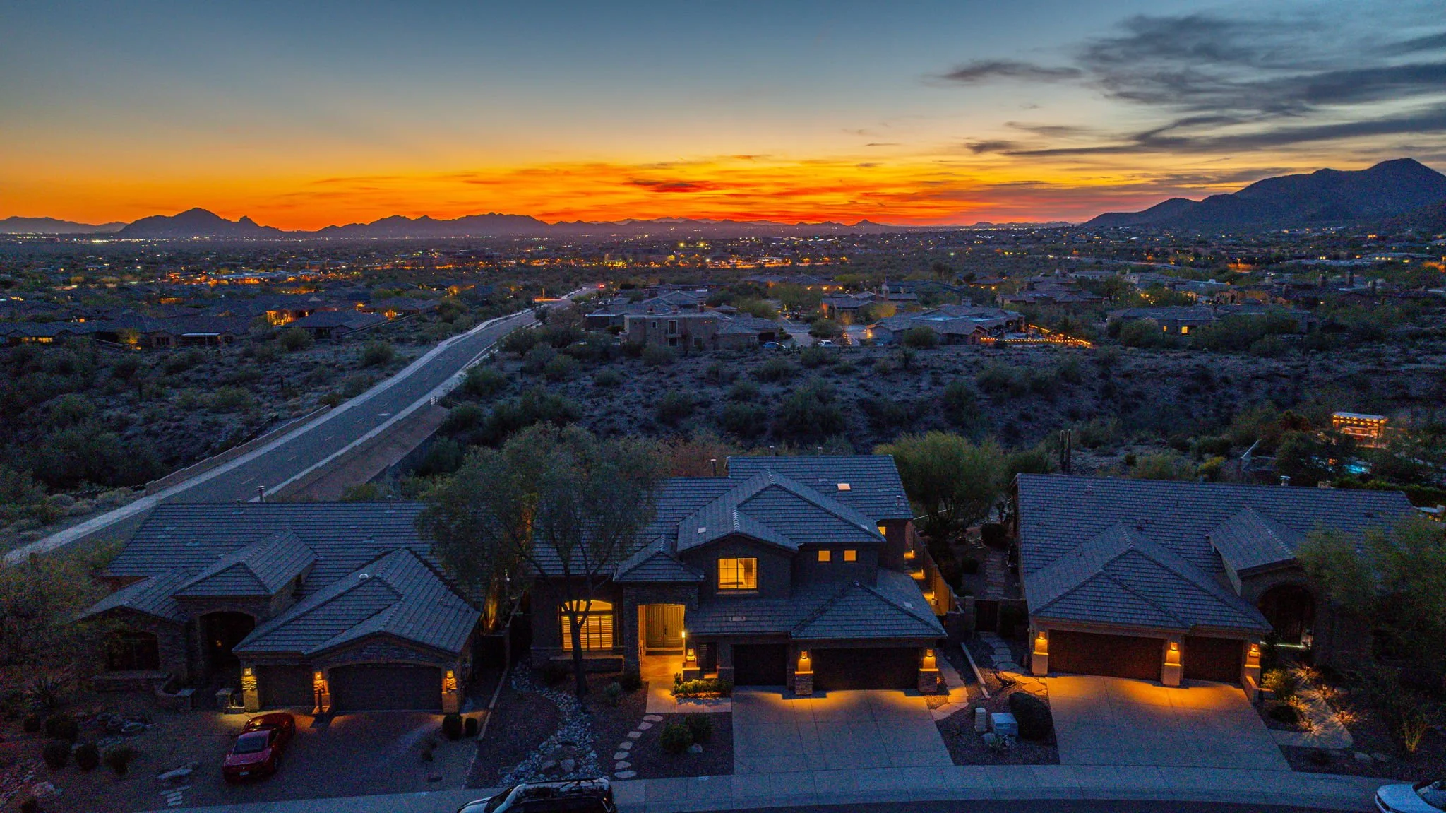 Aerial view of a suburban neighborhood at sunset with mountains in the background, houses with dark roofs, illuminated windows, and cars parked in driveways.