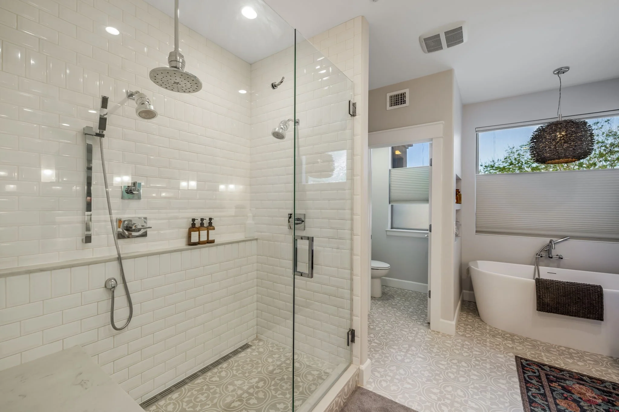 Modern bathroom with a glass-enclosed shower, bathtub, toilet, window, and decorative tile flooring.