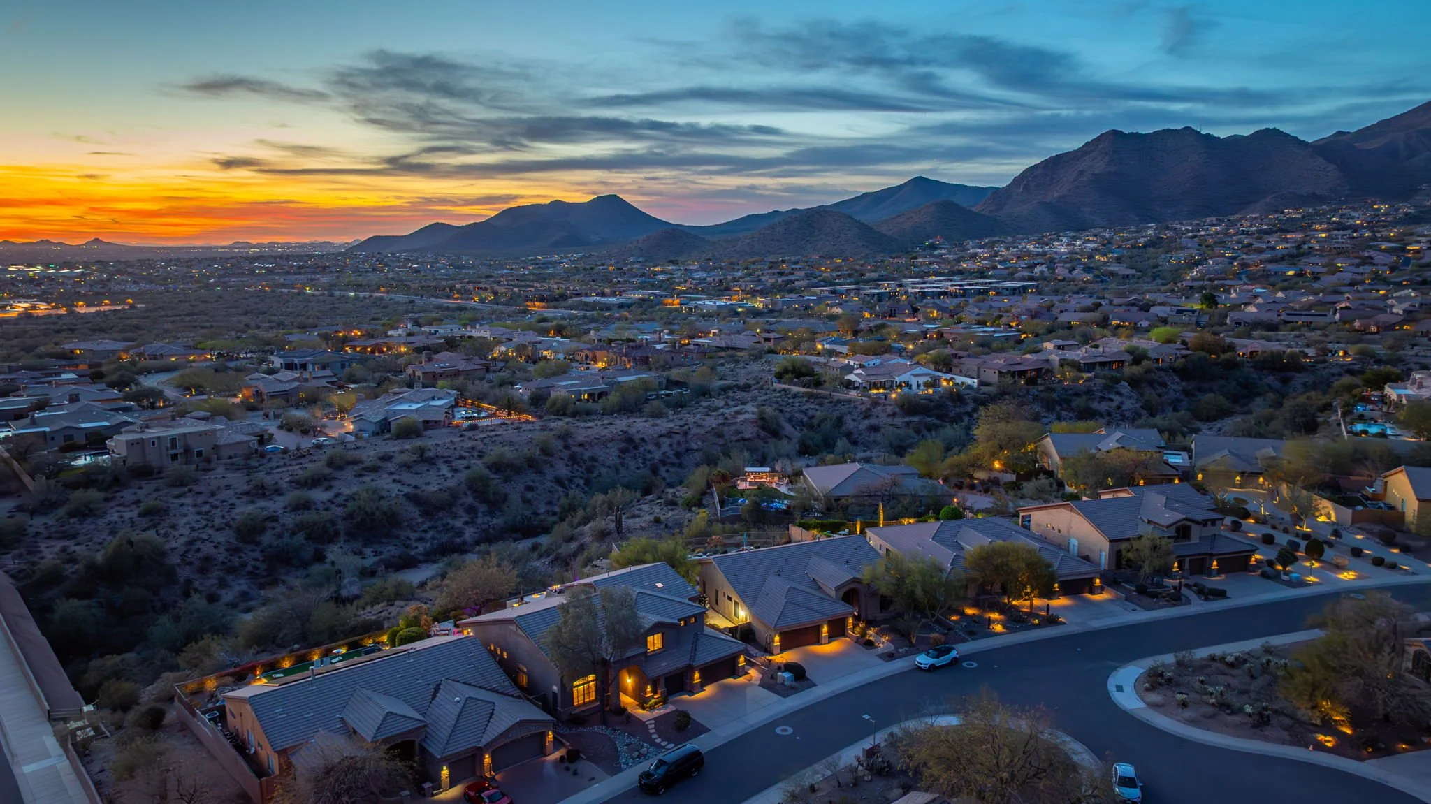 Aerial view of a residential neighborhood at dusk with houses, streets, and mountain range in the background.