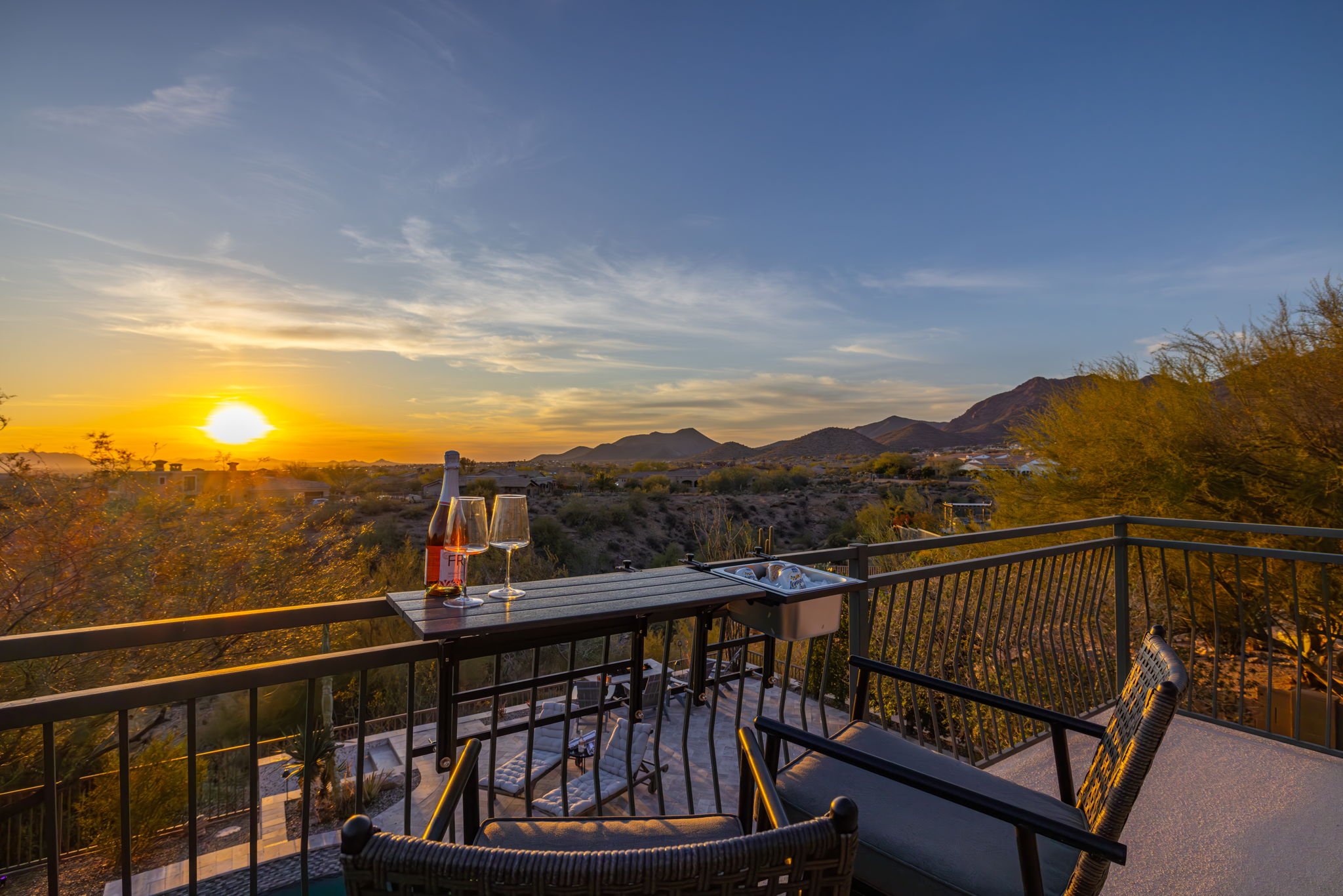 Sunset view from a balcony with wine glasses, a bottle, and outdoor chairs overlooking a desert landscape with mountains in the distance.