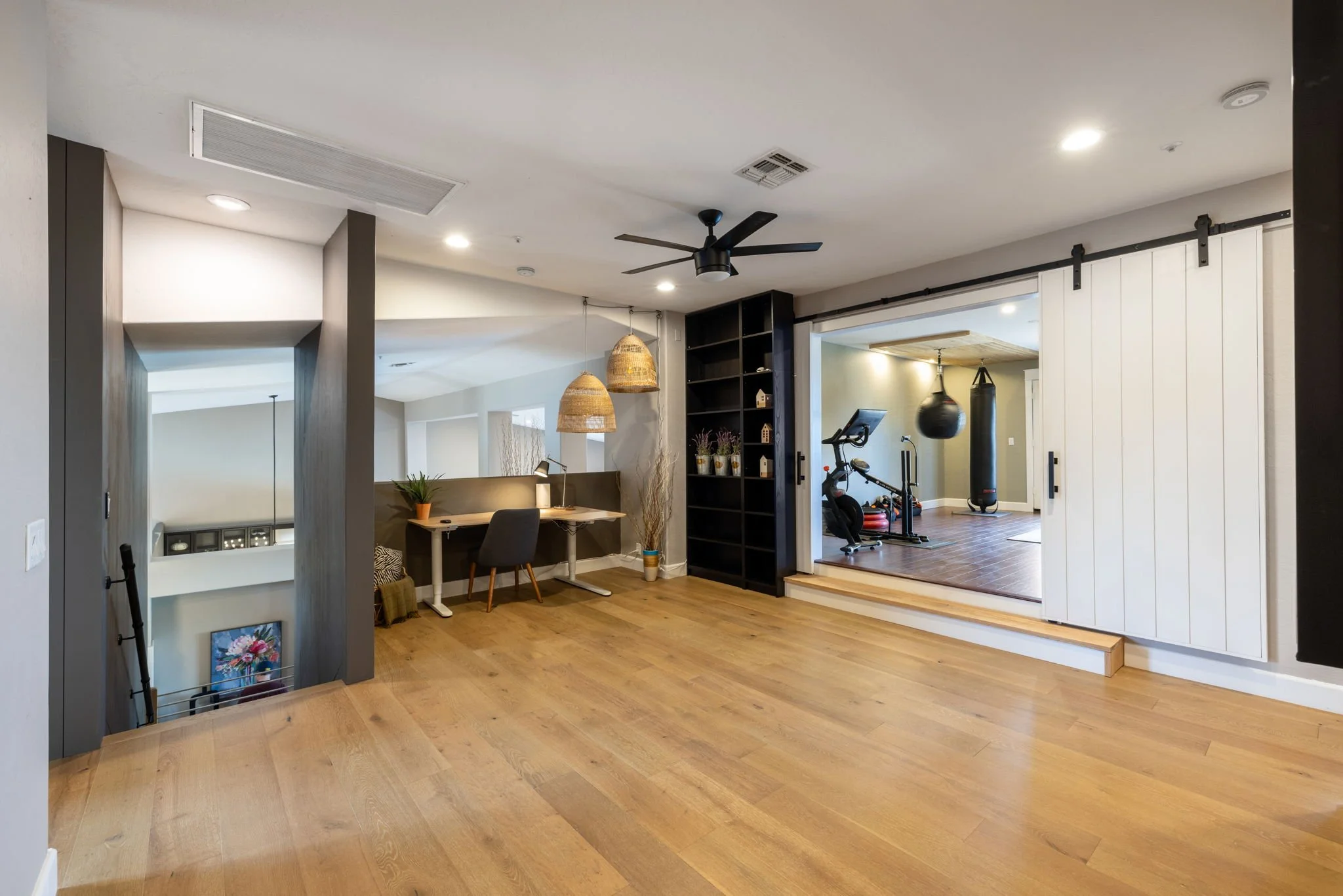Open living space with a wooden floor, a black ceiling fan, a small desk with a chair, pendant lights, and a separate home gym area with punching bag, punching mitts, and exercise equipment.