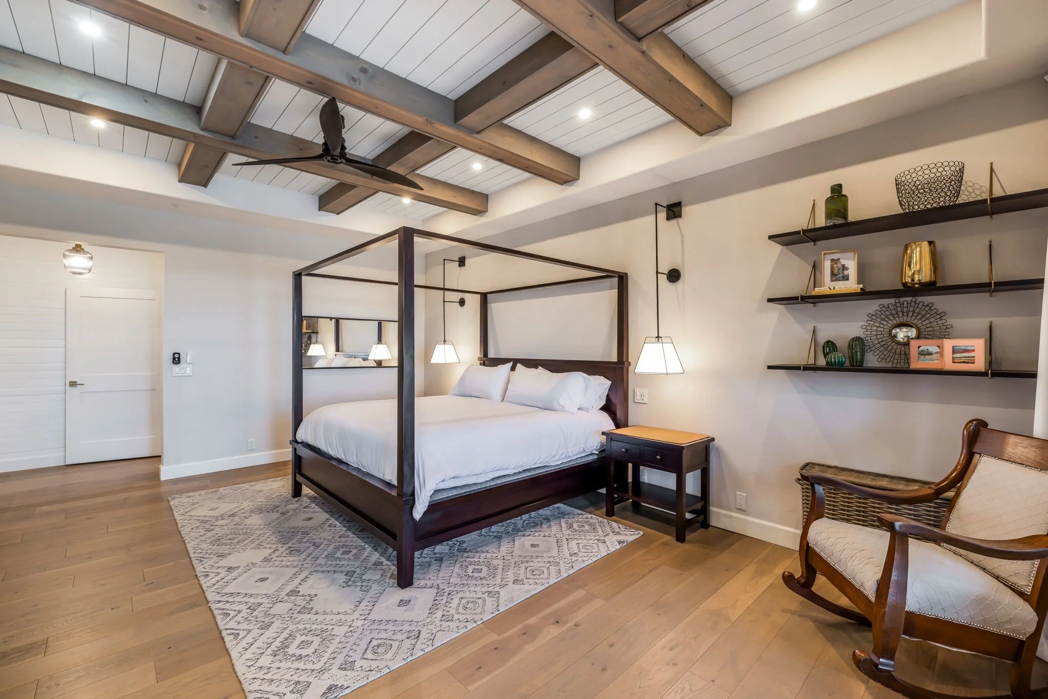 A bedroom with a dark wooden canopy bed, white bedding, and hanging white lamps. There are wood shelves with decorative items on the wall and a wooden rocking chair.