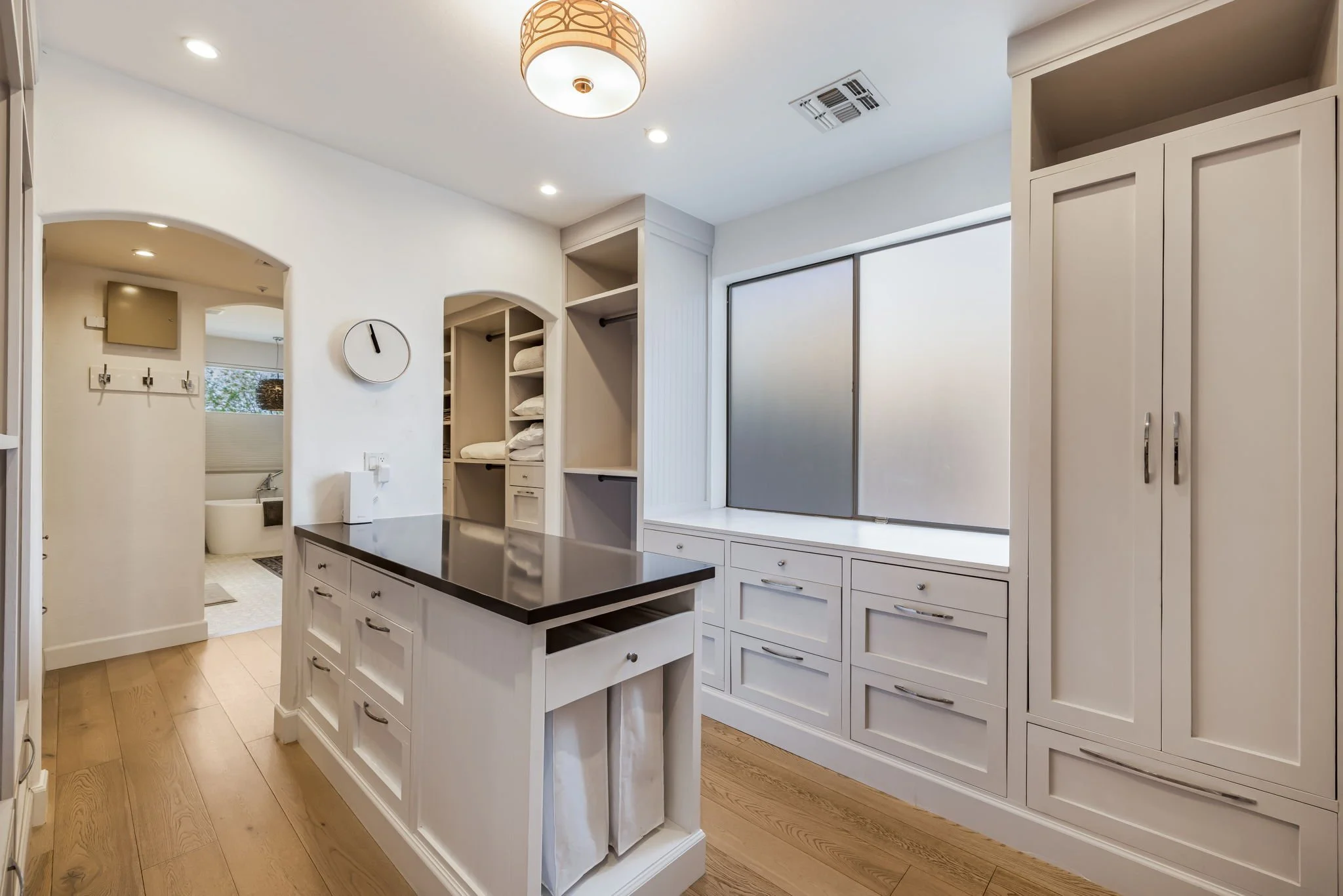 A bright kitchen with white cabinets, a window with gray panels, a wooden island with a black countertop, hardwood floors, and a round ceiling light.