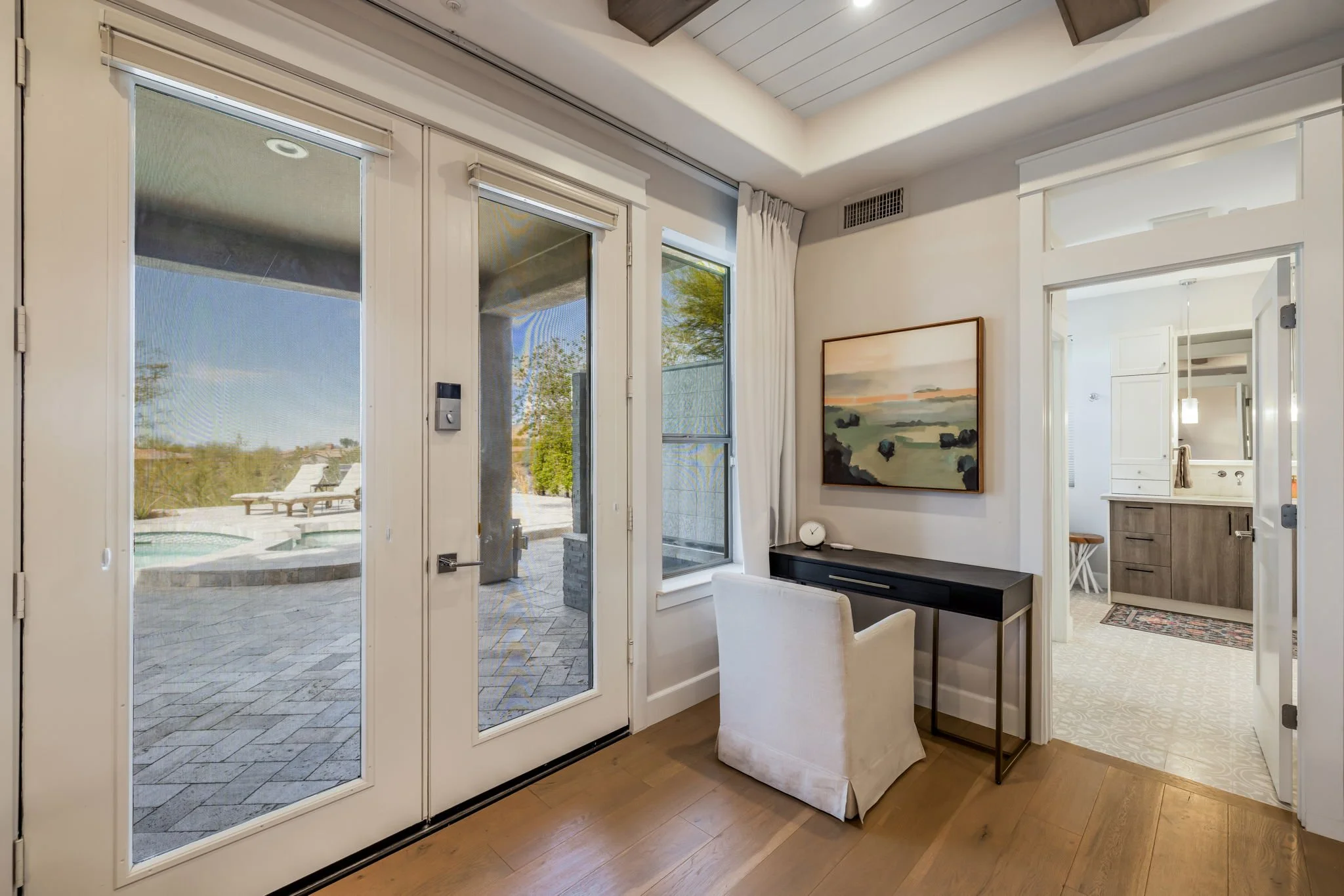 Interior of a room with a view of a patio and hot tub outside, a black desk with a white upholstered chair, a framed landscape painting on the wall, and a door leading to a bathroom with a vanity and mirror.