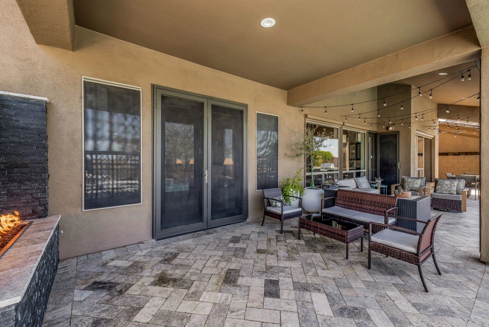 Covered outdoor patio with string lights, patio furniture including chairs and a sofa, potted plants, a fireplace, and large windows and doors on the beige stucco house wall.