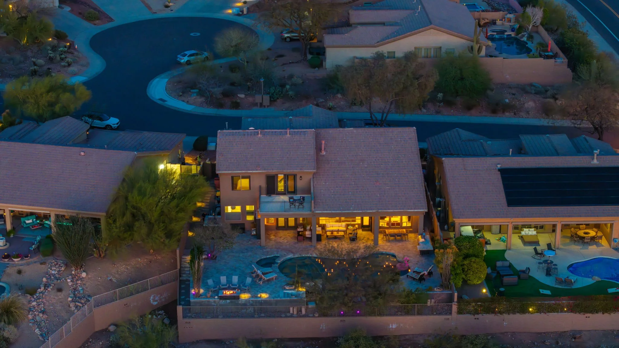 Aerial view of residential area at dusk, showcasing multiple houses with illuminated interiors, a backyard with a pond, lounge chairs, and a fire pit, with mountains and a winding road in the background.