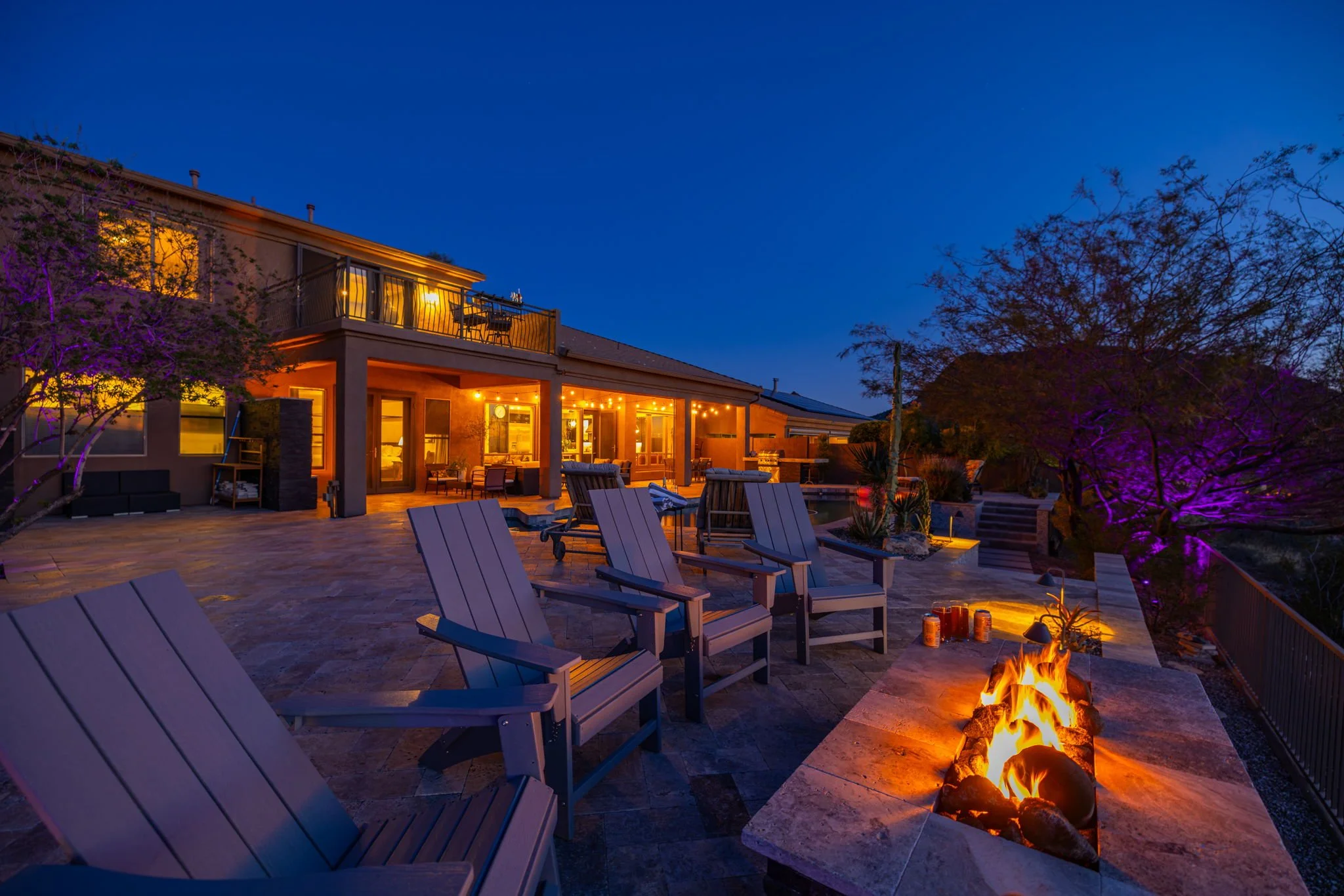 A backyard patio at dusk with chairs around a fire pit, string lights on the house, and trees illuminated in purple.