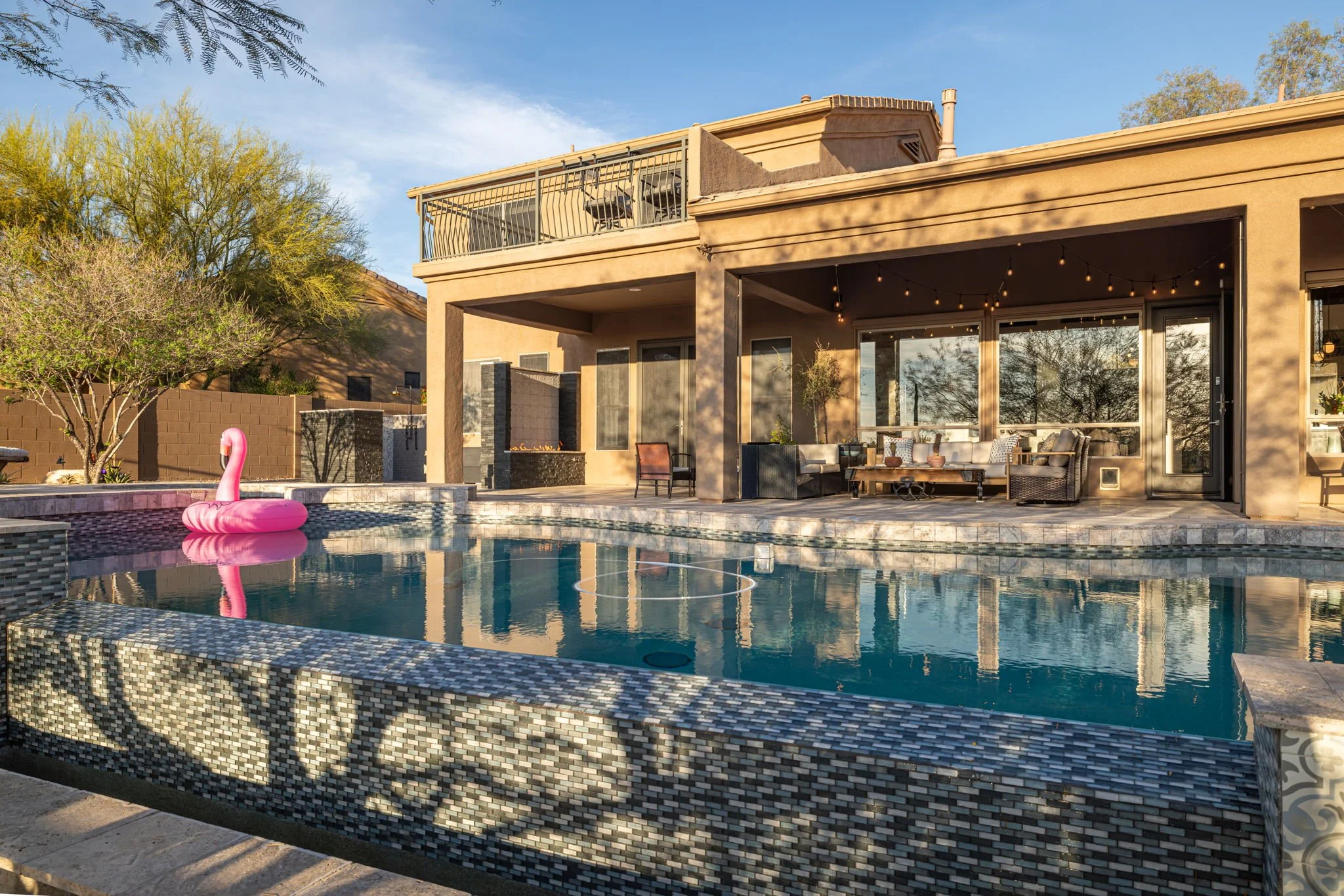 A backyard with a swimming pool, pink inflatable flamingo, patio furniture, string lights, and a house with a balcony and large windows during sunset.
