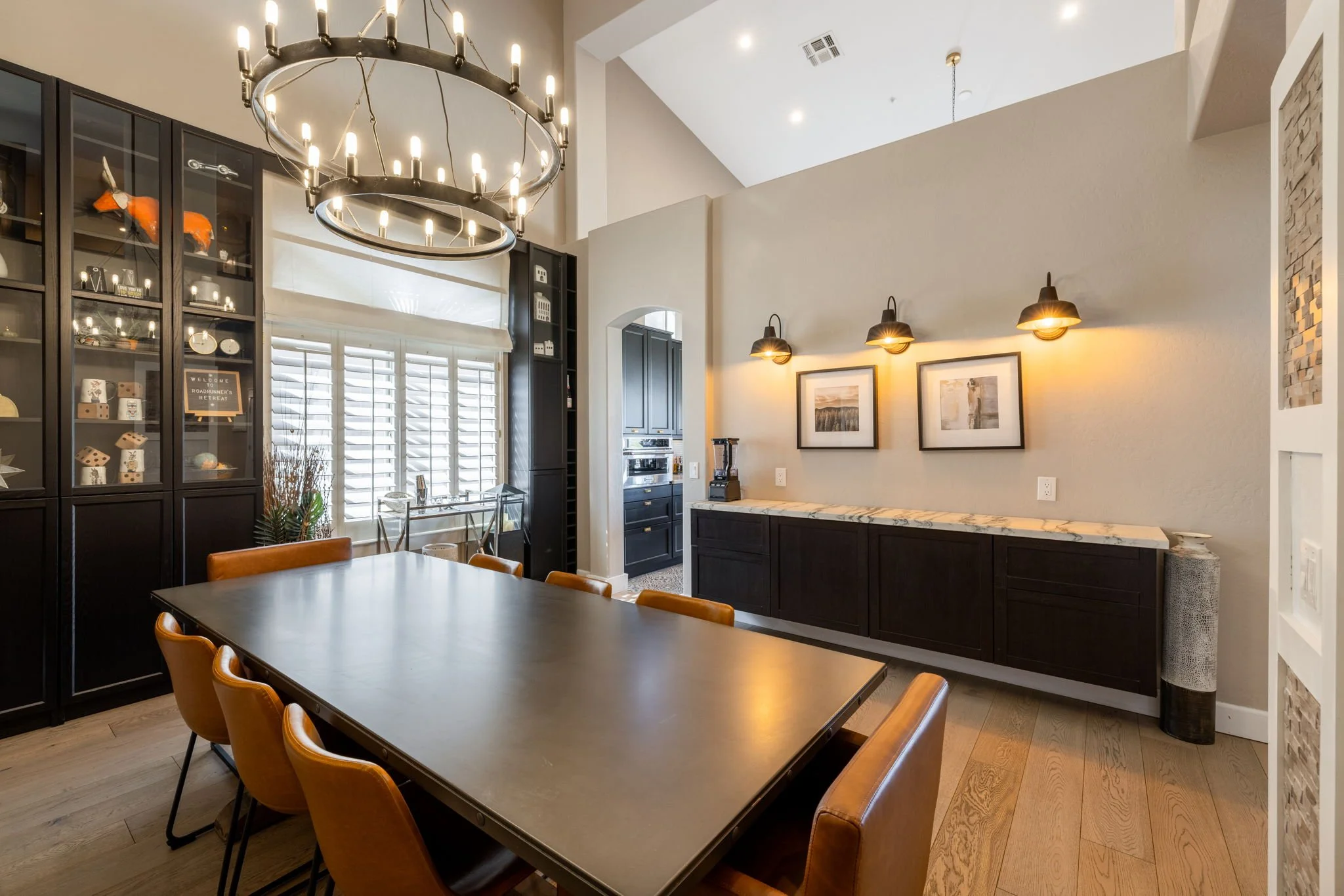 Modern dining room with a large rectangle table, tan leather chairs, black display cabinet, and a sideboard with framed artwork above it, illuminated by wall sconces and a large chandelier.
