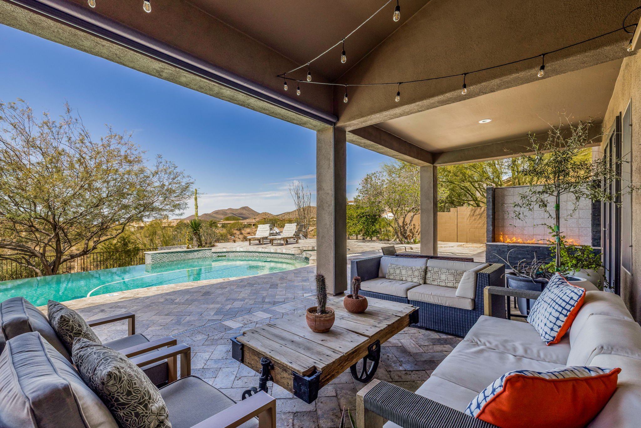 Covered patio with outdoor seating, potted cacti, a fire feature, and a view of a swimming pool and desert landscape in the background.