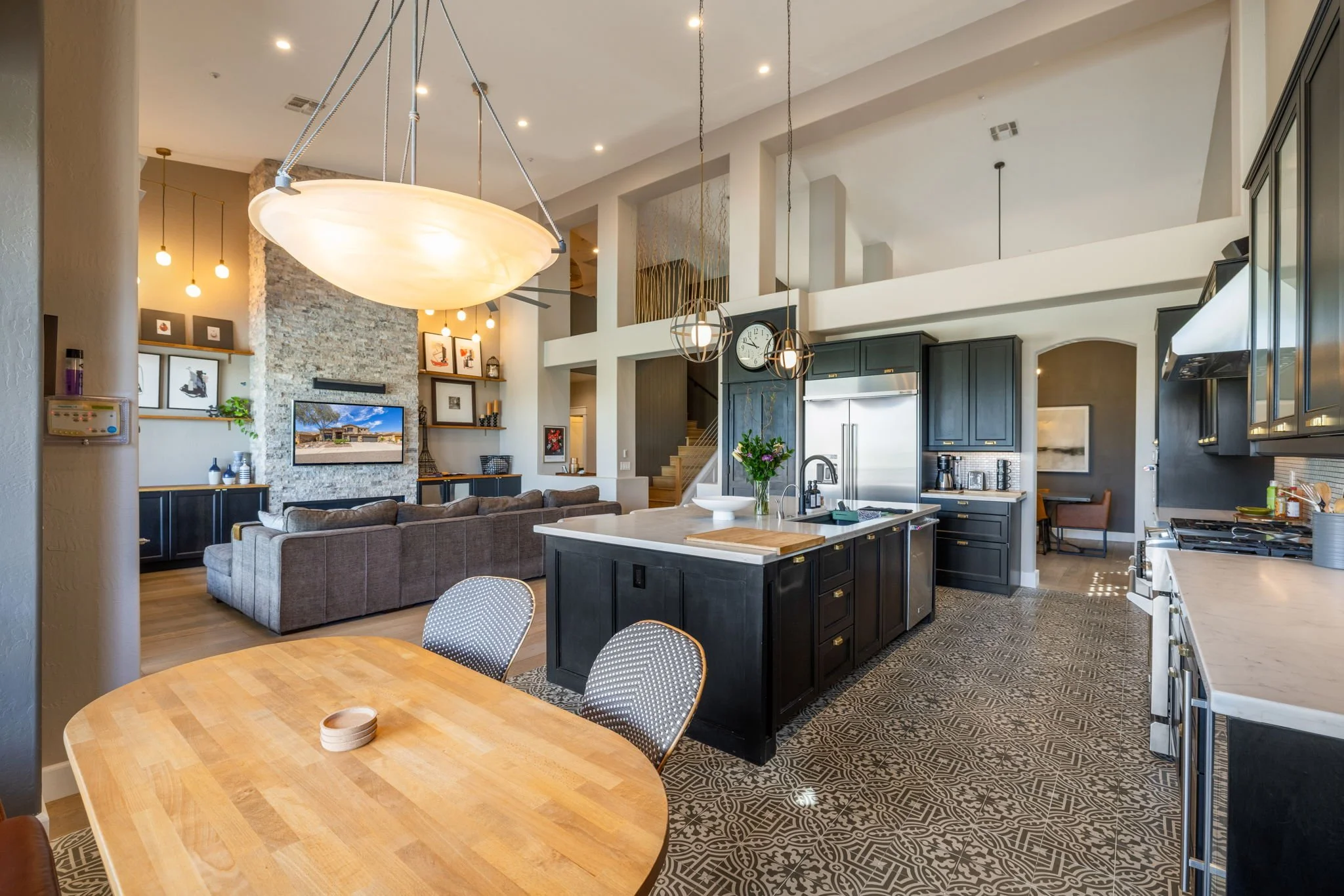 Open-concept living room and kitchen with modern decor, a large gray sofa, a stone fireplace with a wall-mounted TV, black kitchen cabinets, a kitchen island with a sink and flowers, patterned floor tiles, and pendant lighting.