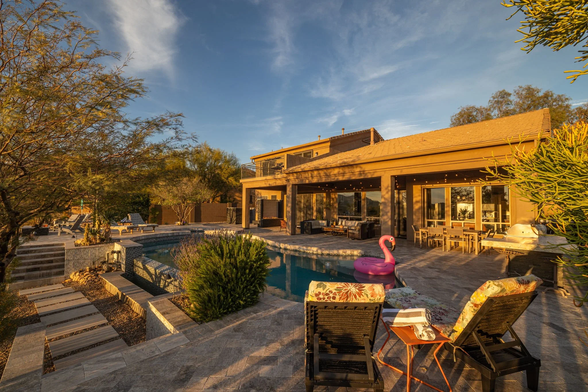 Backyard of a house with a swimming pool, lounge chairs, a pink flamingo pool float, surrounded by trees and patio furniture, during sunset.