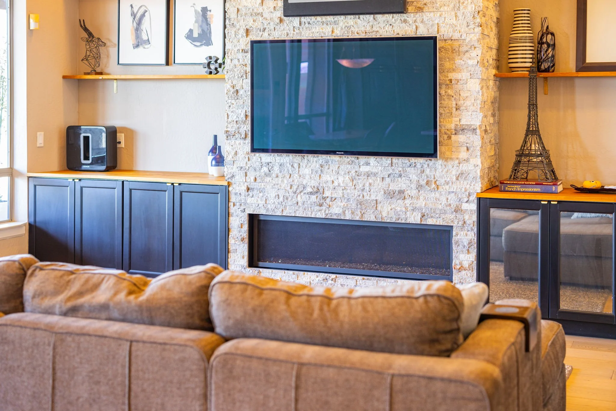 Living room with beige sofa, TV mounted on a stone fireplace, black cabinets, decorative vases, and wall art.